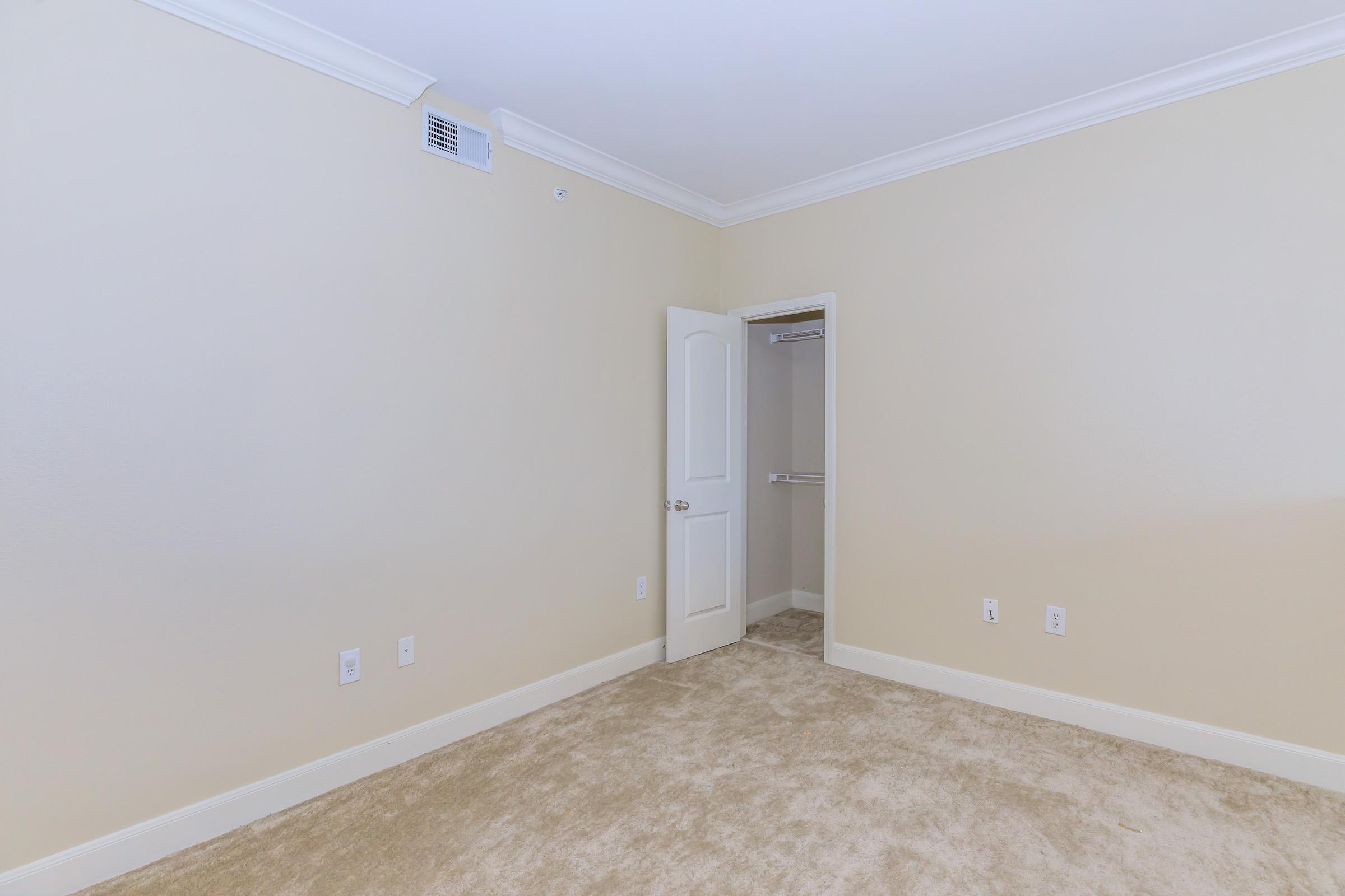 A spacious, empty room with beige walls and light carpeting. There is a white door leading to a closet on the right, and the room has a simple crown molding along the ceiling. The overall look is bright and minimalistic, suitable for a new setup or decoration.