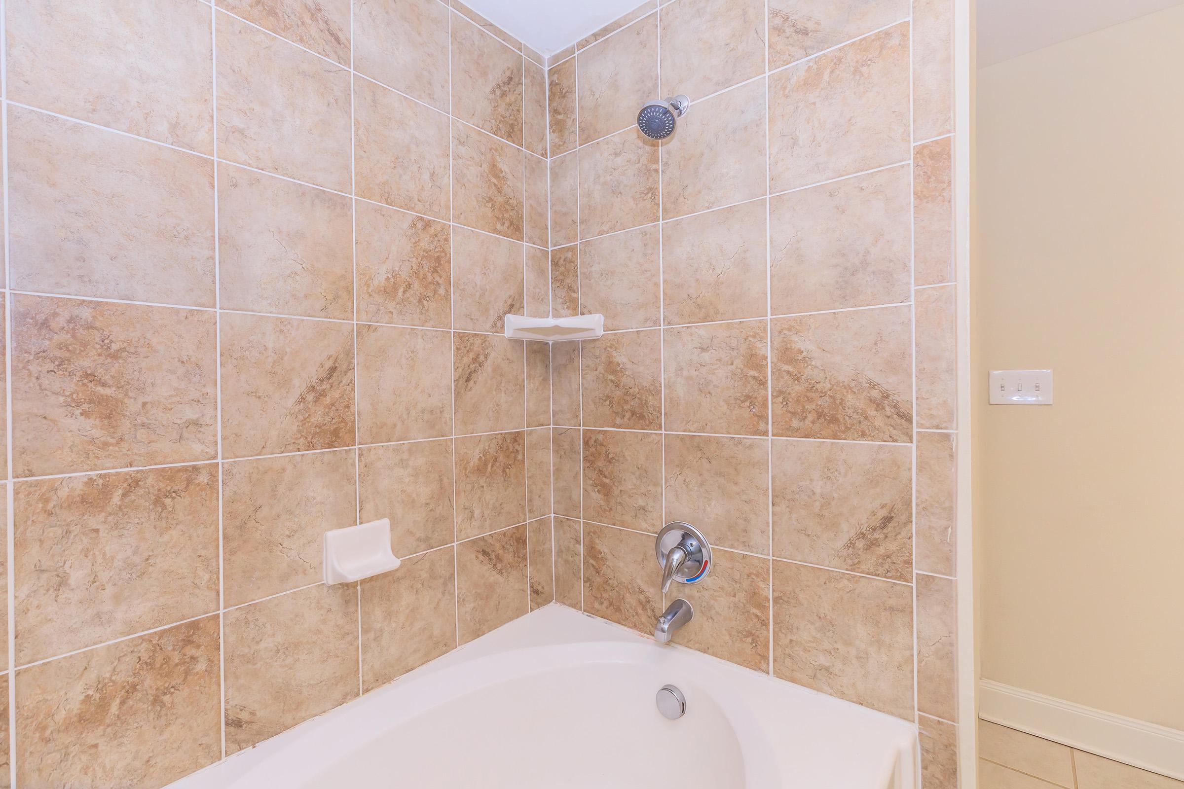A corner shower area featuring beige and brown tiled walls, a chrome showerhead, and a white bathtub. A small shelf is installed on the wall, and a light-colored wall is visible beside the shower area.