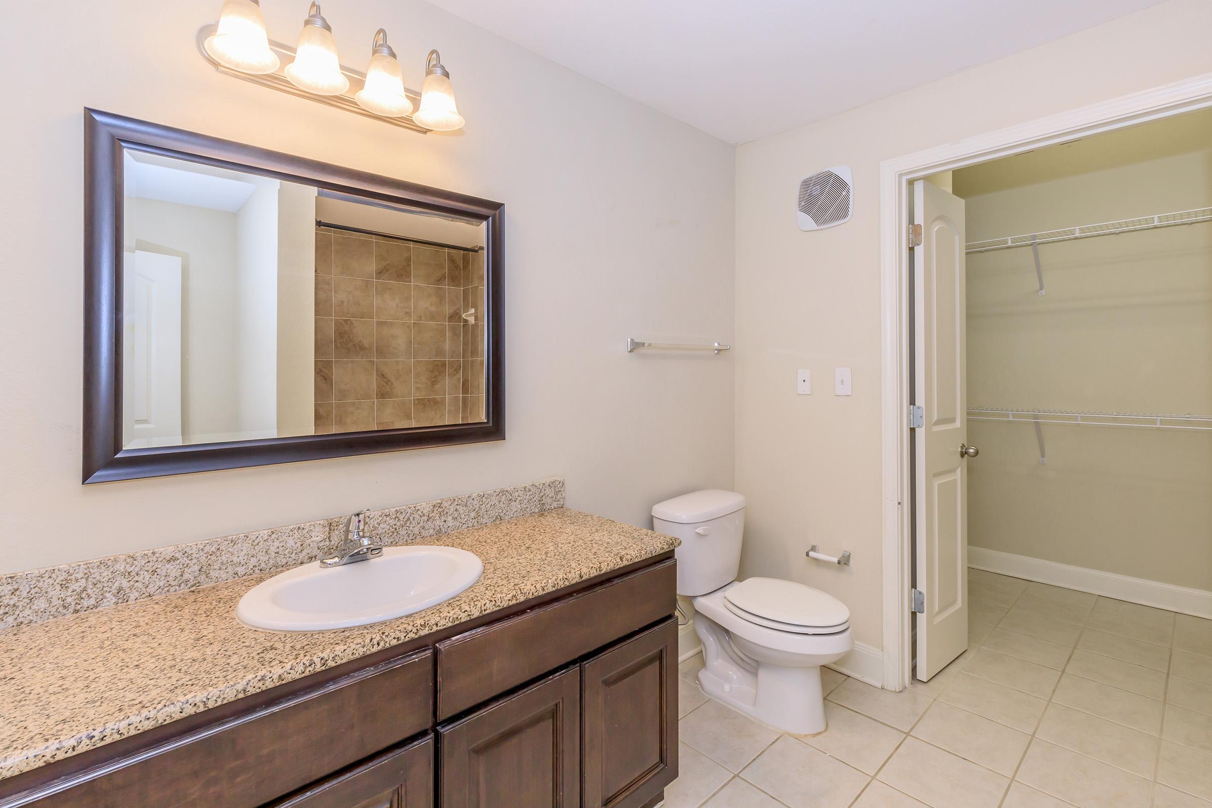 A modern bathroom featuring a granite countertop with a sink and dark wood cabinetry, a white toilet, and a large mirror above the sink. Beige tiled walls surround a shower area, and there's a spacious closet with a white door on the right. Bright lighting enhances the clean, contemporary look.
