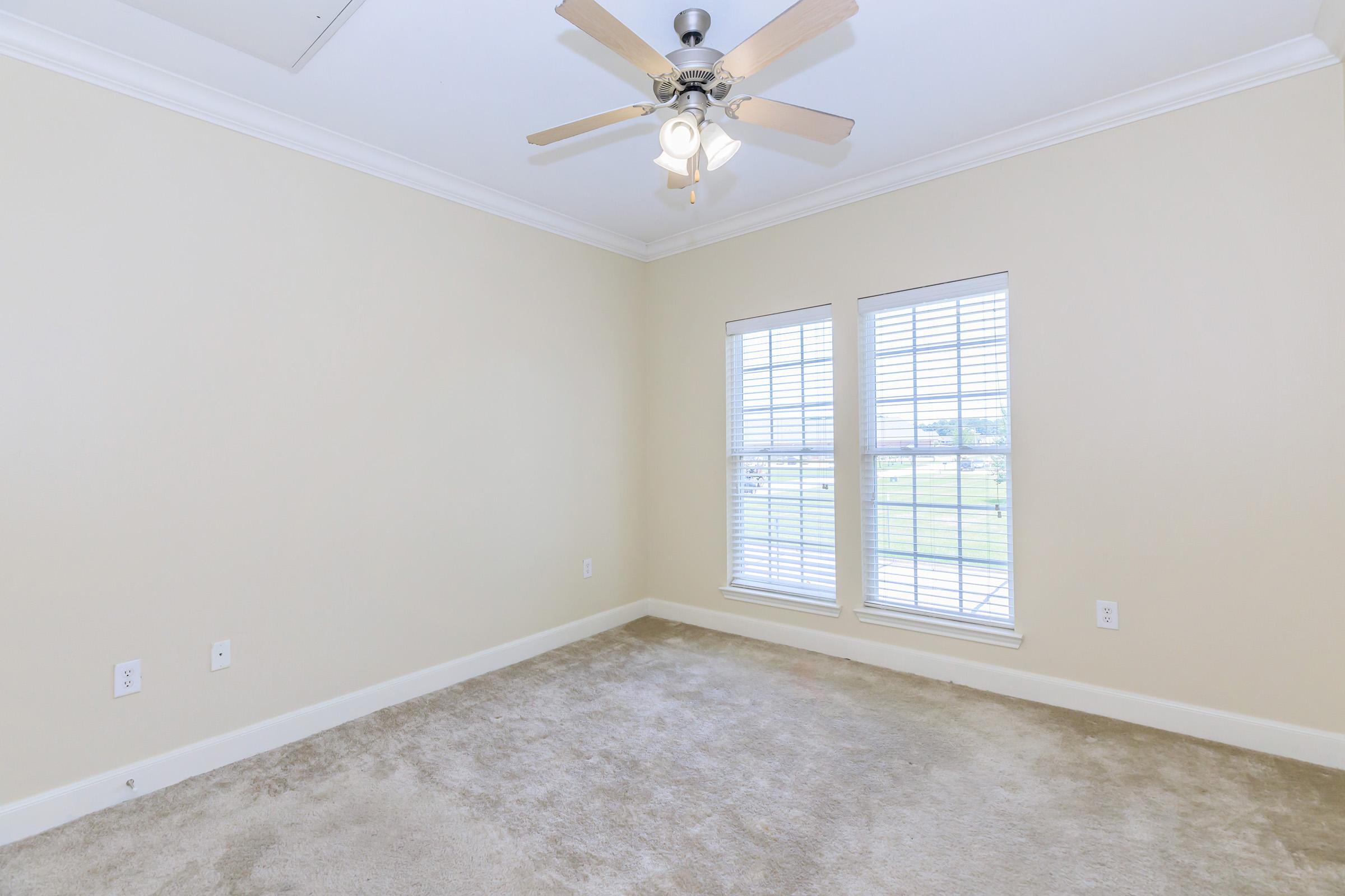 A clean, empty room with light beige walls and a light carpet. The room features a ceiling fan with wooden blades and two windows with white blinds, allowing natural light to fill the space. The floor is clear of furniture, creating a blank canvas feel.