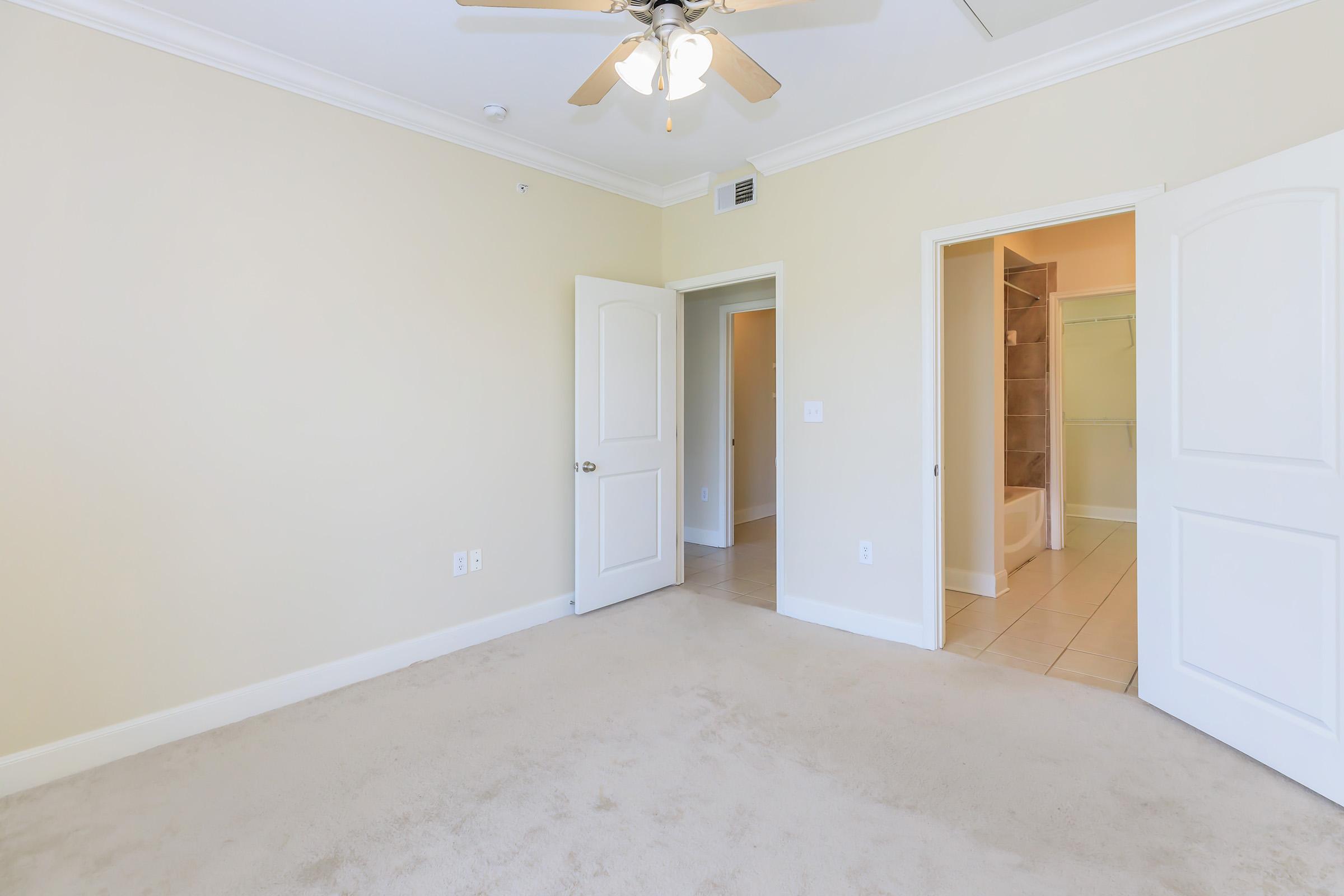 A bright, empty room featuring beige walls and a ceiling fan. The room has a light carpeted floor and two doors: one leading to a bathroom with a shower, and another door open to a hallway. Natural light fills the space, creating an airy atmosphere.