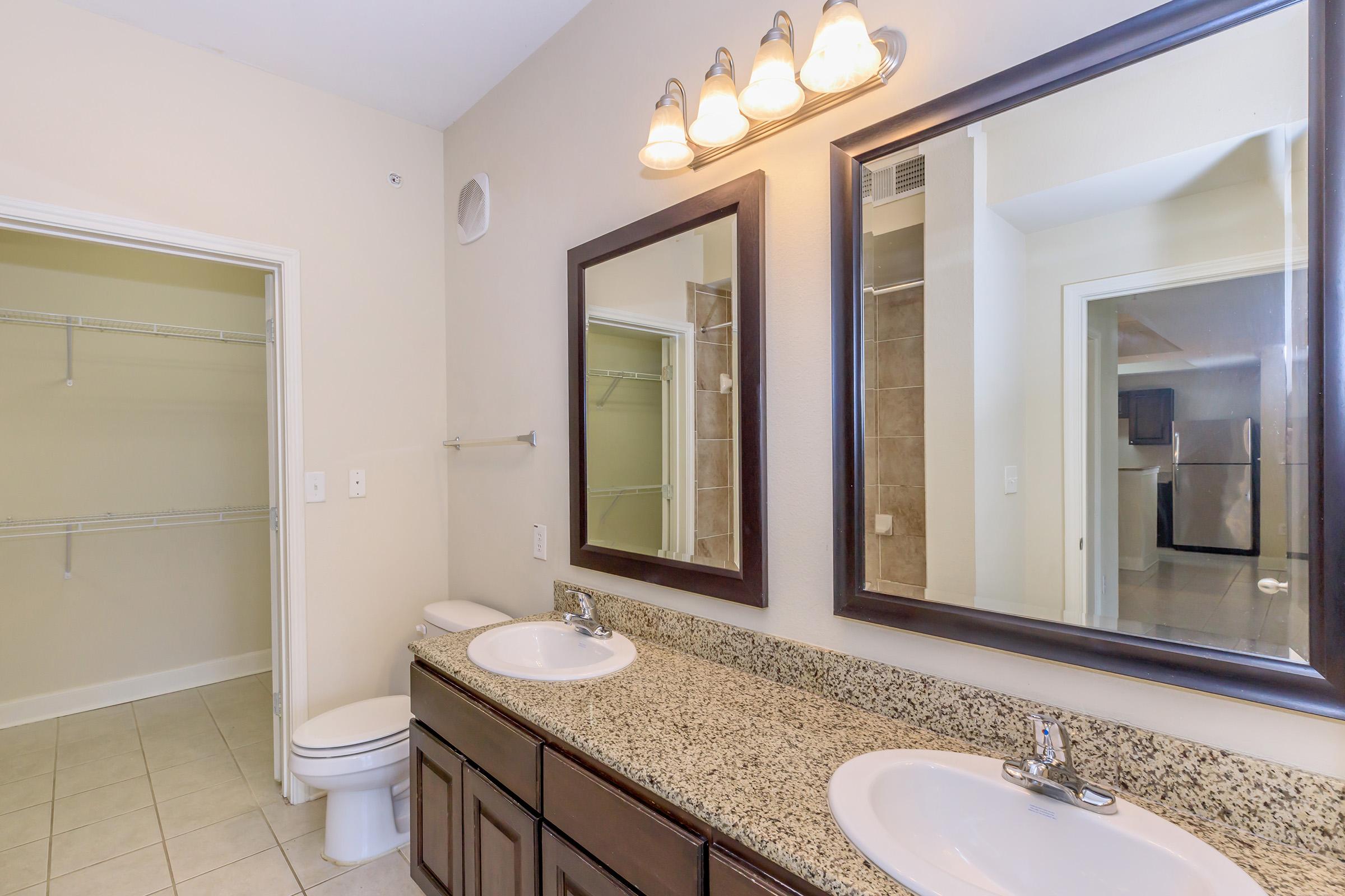 Dual vanity bathroom featuring a stone countertop, two sinks, and large mirrors. Well-lit with wall-mounted fixtures. A linen closet is visible on the left, and the room leads to a space with tiled flooring and an adjoining area with kitchen appliances in the background.