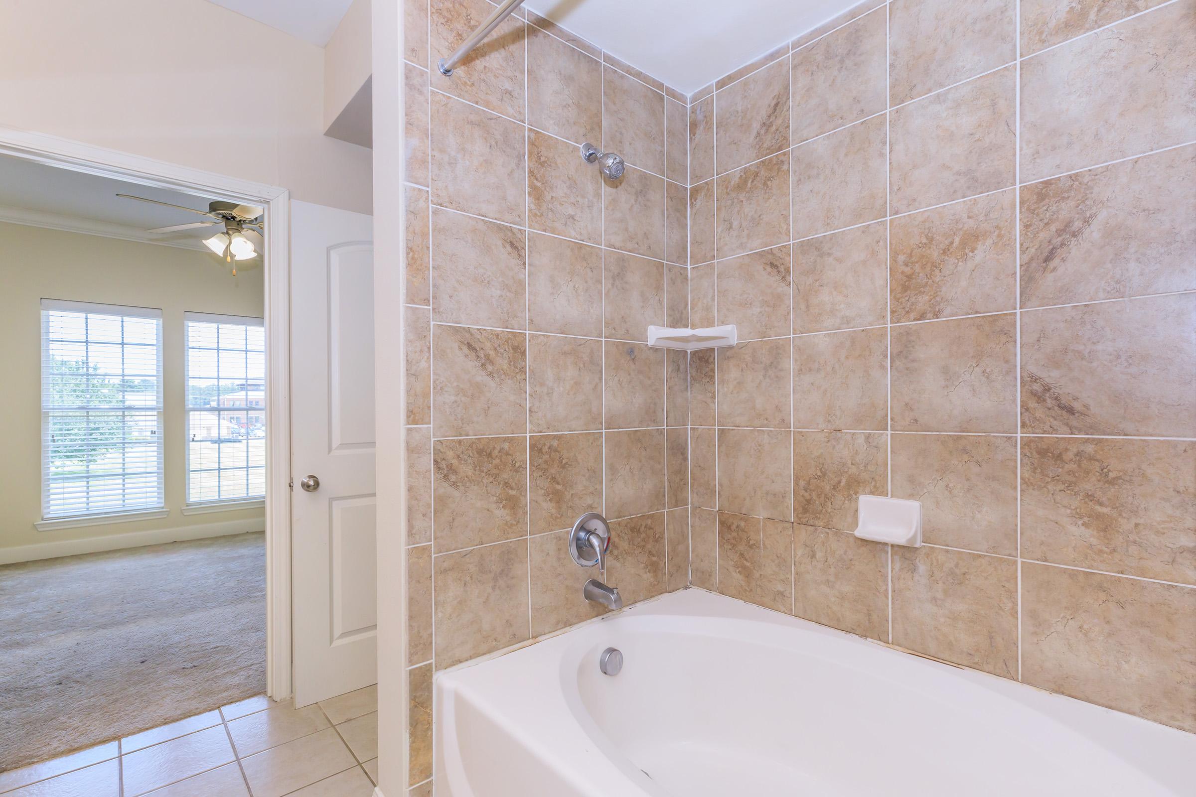 A modern bathroom featuring a bathtub with a showerhead, tiled walls in warm beige tones, and a white soap dish. The background shows an open doorway leading to a room with a ceiling fan and large windows, allowing natural light to enter.
