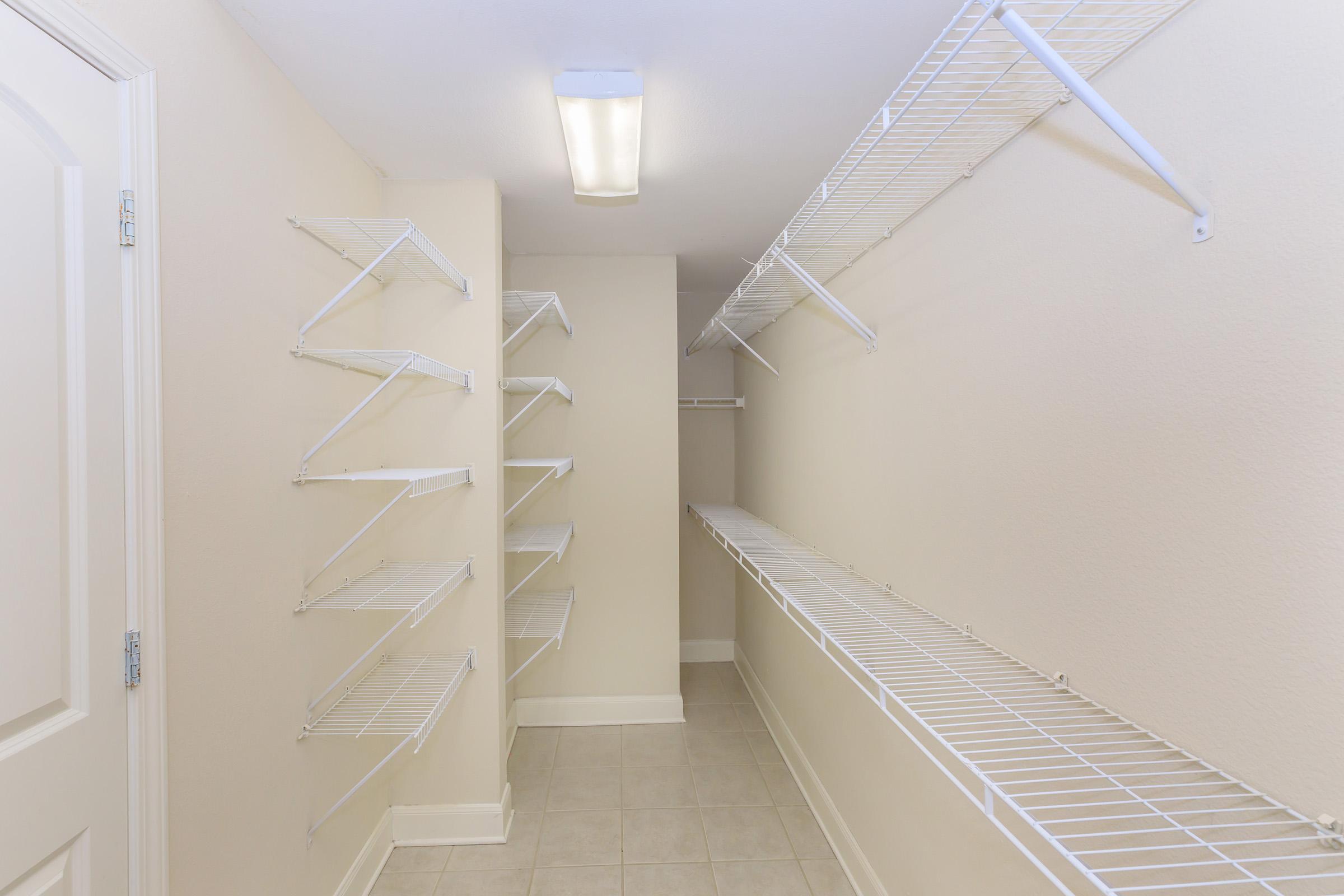 Empty walk-in closet with white wire shelving along the walls and a long shelf at the back. The walls are painted a light beige, and the flooring is tiled. A ceiling light fixture illuminates the space.