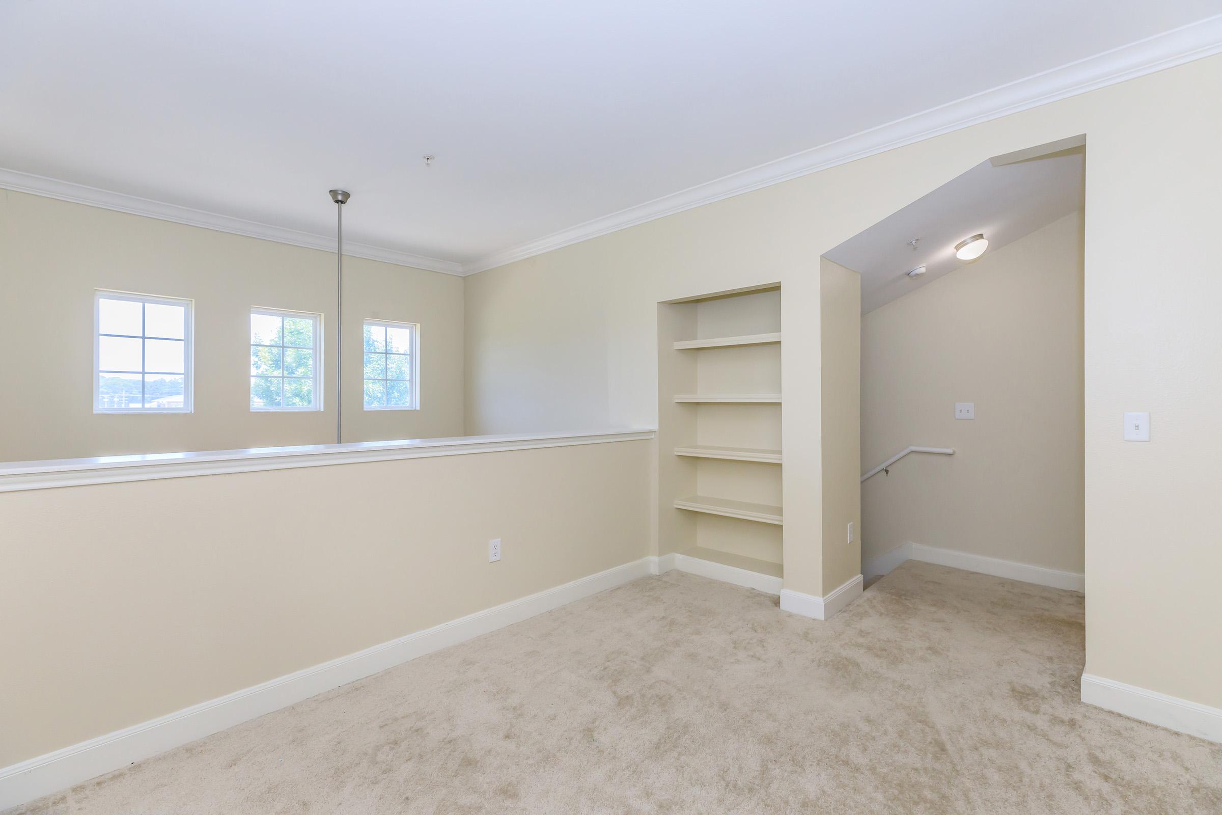 A spacious room with light-colored walls and carpet, featuring three large windows allowing natural light. There is a built-in bookshelf along one wall, and a staircase leading to another area is visible on the right. The space is clean and well-lit, creating an inviting atmosphere.