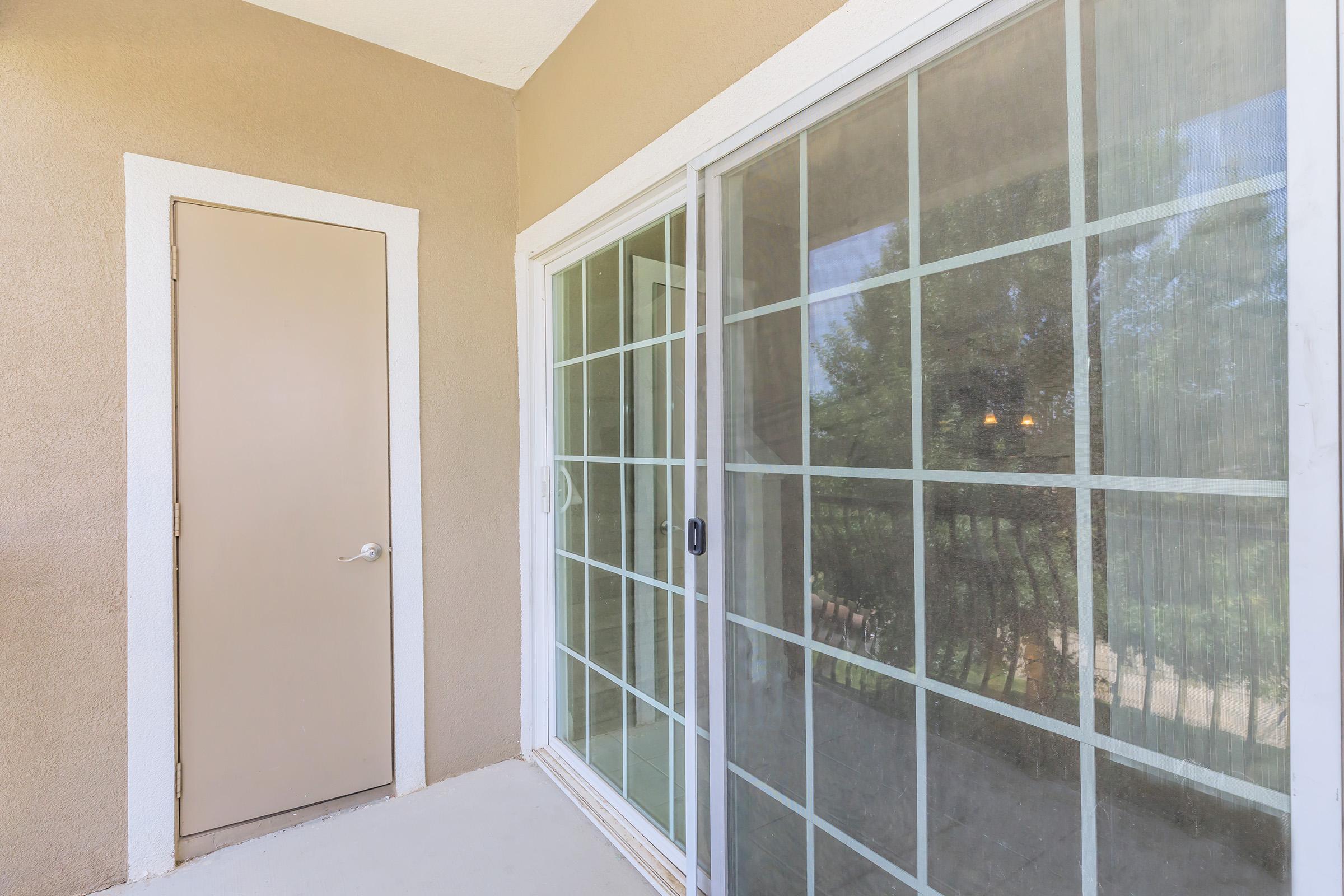 A view of a balcony area featuring a door leading to a room and a large sliding glass door with grid patterns. There is a light beige wall surrounding the doors, and natural light is visible through the glass, providing a glimpse of the outdoors.