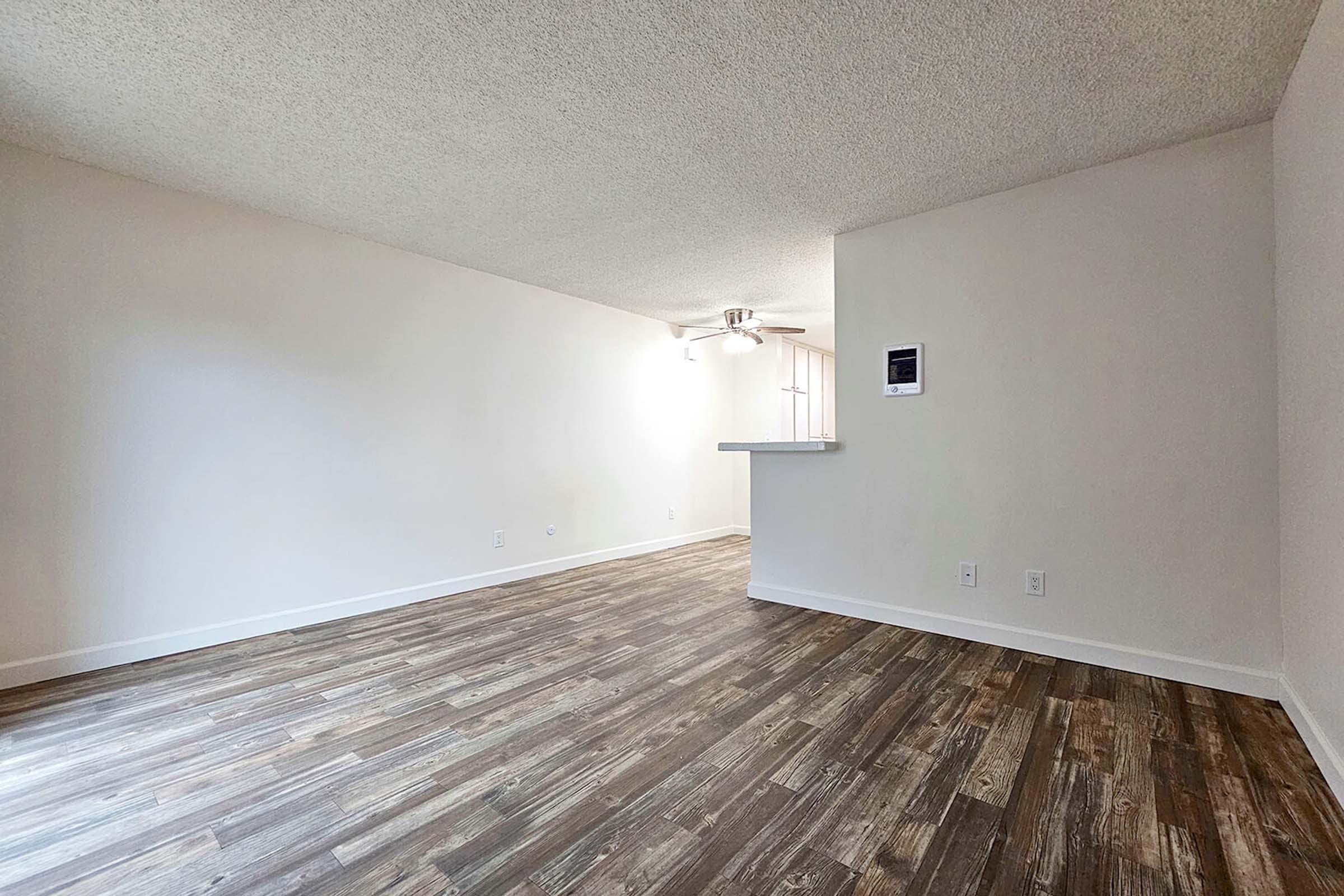 A spacious, empty room with light-colored walls and a textured ceiling. The floor is covered in wide wooden planks. There is a ceiling fan and a small bar area leading to a kitchen, with natural light streaming in from a window. The overall atmosphere is airy and ready for furnishing.