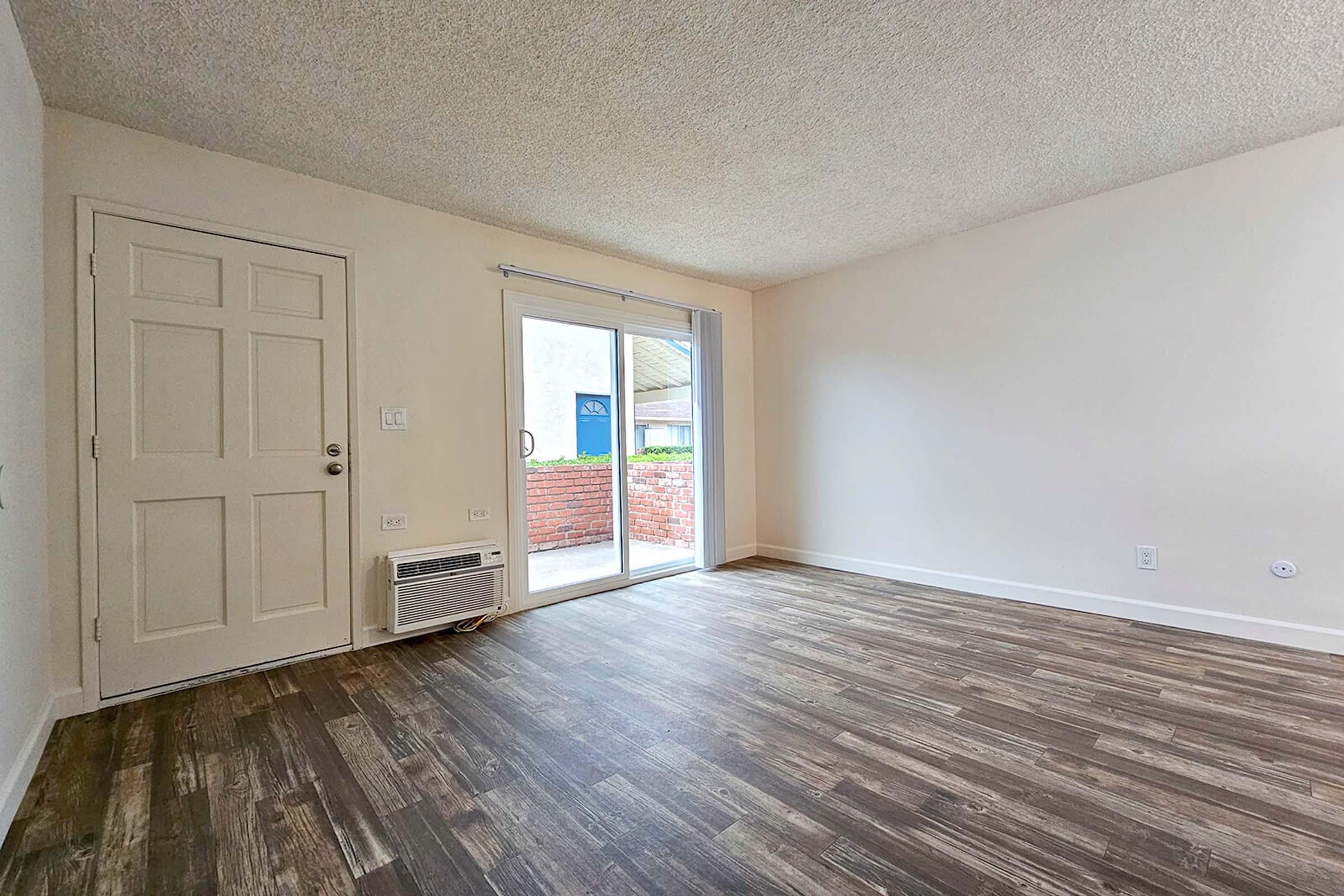 A spacious, empty room with light-colored walls and a textured ceiling. There is a door leading to a patio area, and a large window allows natural light to enter. The floor is covered with dark wood-like laminate flooring, and there is an air conditioning unit on the wall near the door.