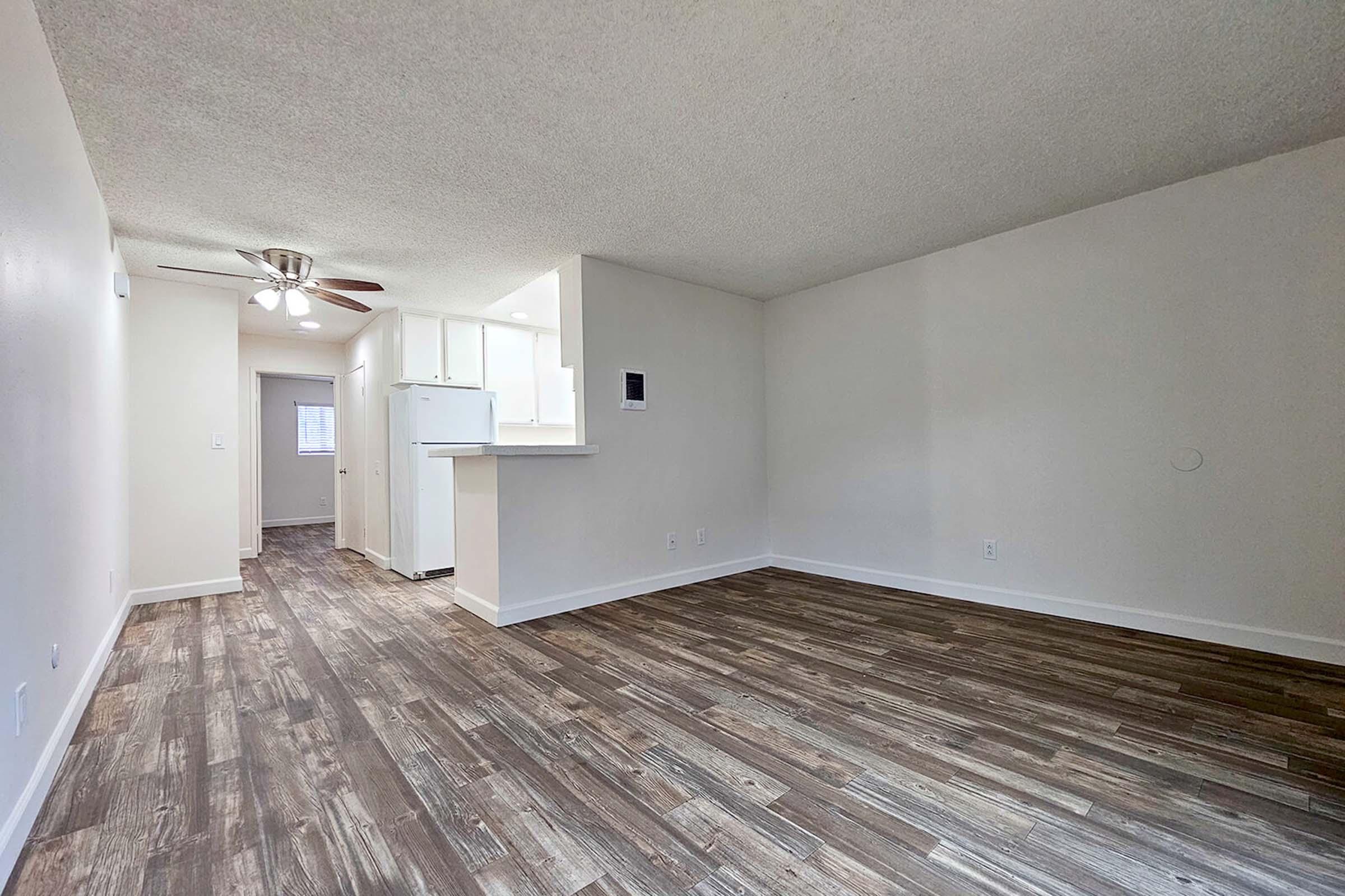 An empty room with beige walls and a textured ceiling. It features a wooden floor, a ceiling fan, and a kitchen area with a white refrigerator. A doorway leads to another room in the background, and the space is well-lit with natural light.