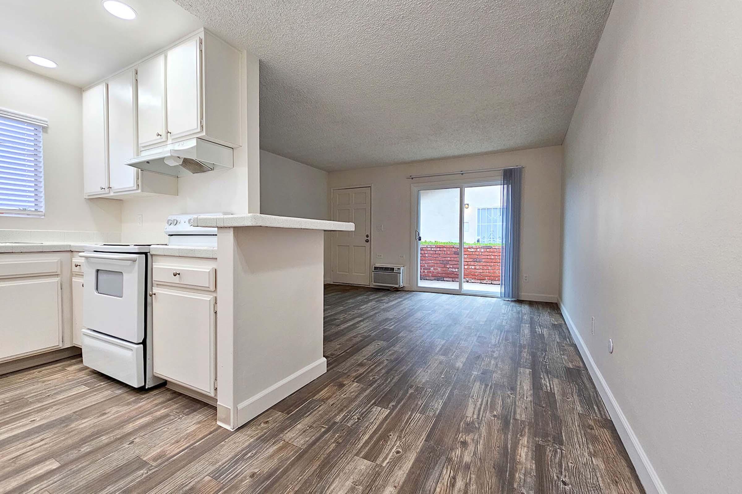 A spacious, updated living area with wooden flooring, featuring a kitchen on the left with white cabinets and an electric stove. To the right, there's a sliding glass door leading to a patio area. The walls are painted in light neutral tones, creating a bright and inviting atmosphere.