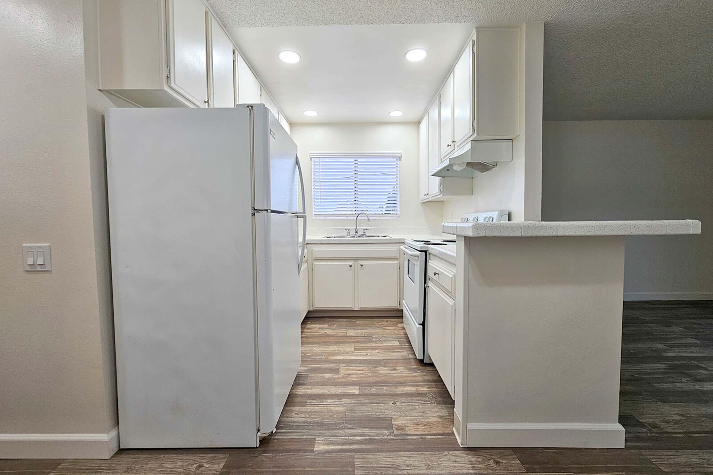 Compact kitchen featuring white cabinetry, a refrigerator, stove, and countertop. Natural light enters through a window above the sink, and modern fixtures add a clean aesthetic. The floor is covered with wood-like tiles, giving the space a warm feel. The simple layout enhances functionality in this small area.