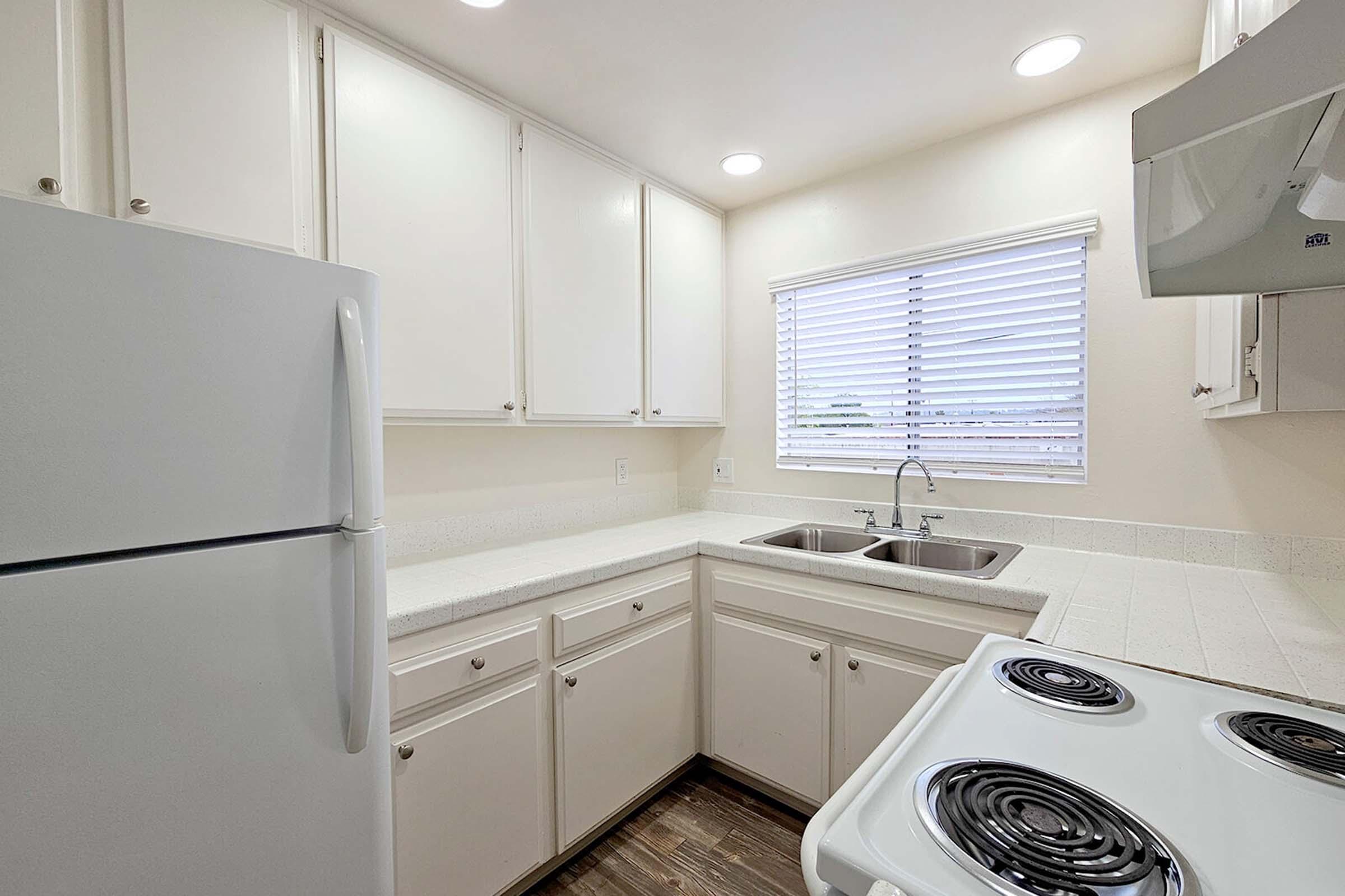 A clean, modern kitchen featuring white cabinets, a double sink, a white refrigerator, and a black coil stove. Natural light streams in through a window with blinds, illuminating the countertop and giving the space a bright, inviting feel.