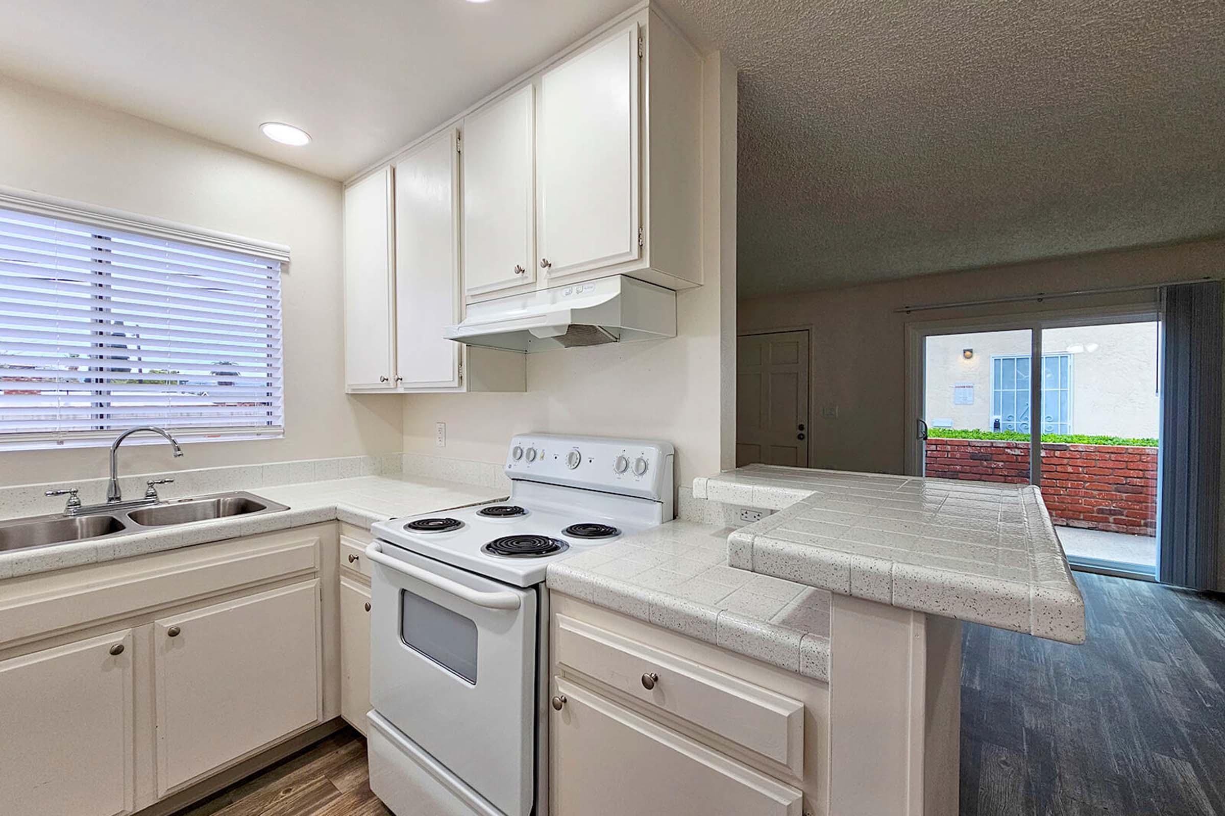 A modern kitchen featuring white cabinets, a stainless steel sink, and a white electric stove. The countertops are light-colored, and there is a window with blinds allowing natural light. A doorway leads to an outdoor space, enhancing the open feel of the area. The flooring is dark wood.