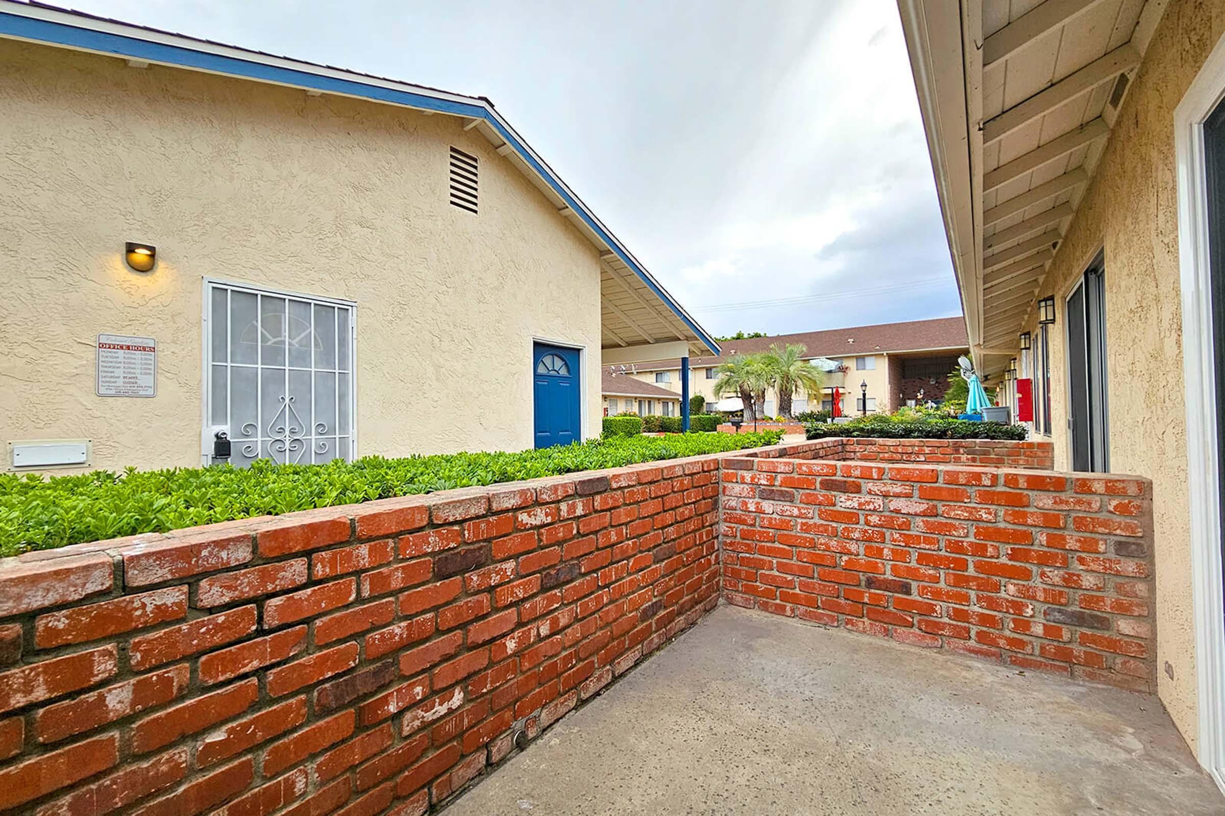 A view of a brick wall in the foreground, leading to a pathway between two beige buildings. One building features a blue door, while another has a white door with decorative ironwork. Green shrubs line the pathway, and there is a statue visible in the background, surrounded by greenery. The sky is overcast.