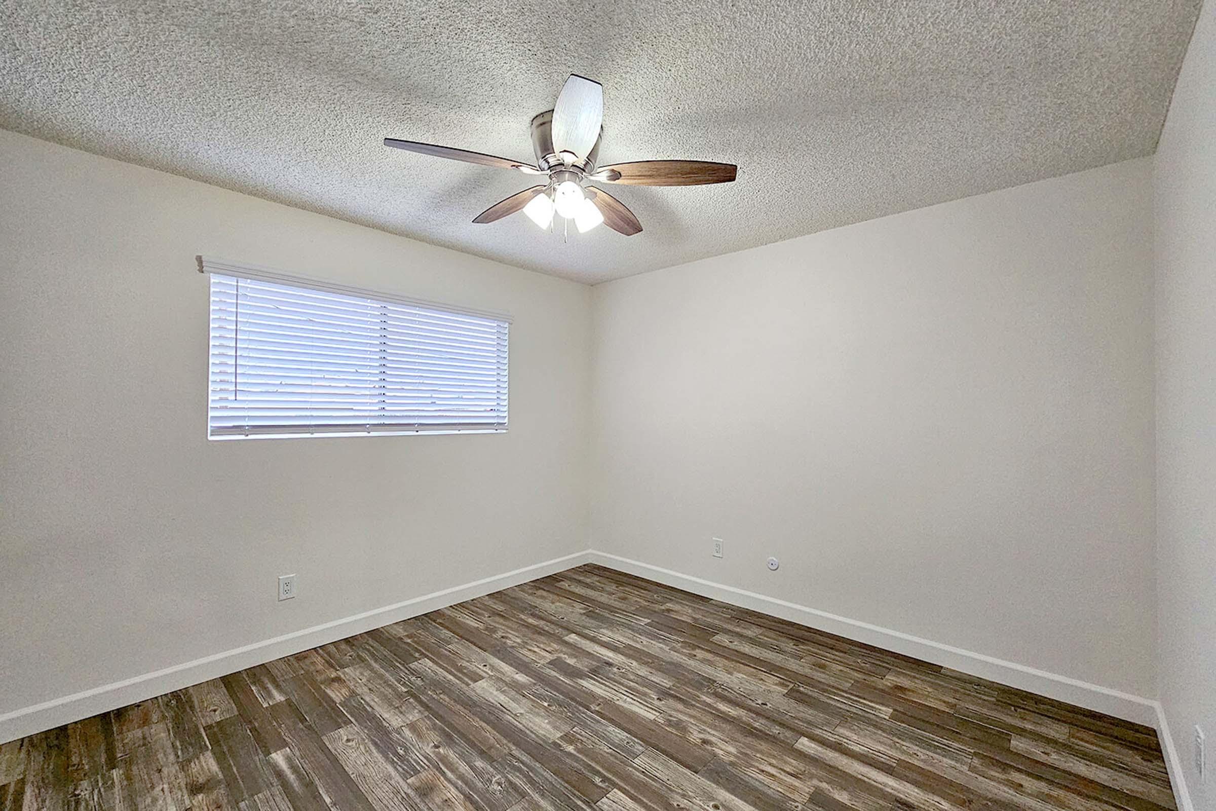 A bright, empty room featuring light-colored walls and a ceiling fan. There is a window with white blinds allowing natural light to enter, and the floor is covered with wooden planks, creating a warm and inviting atmosphere. The room has a spacious feel with no furniture in it.