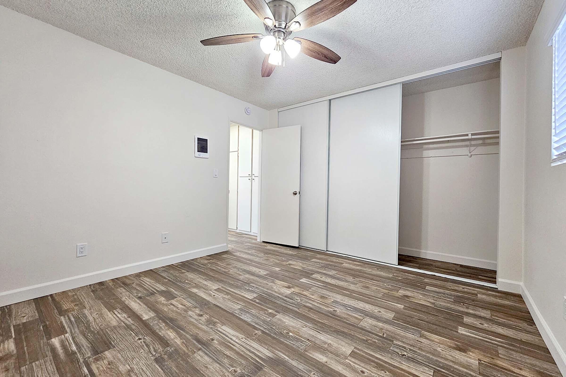 A small, empty room with light-colored walls and wooden flooring. The ceiling features a fan with wooden blades. There's a closed closet with sliding doors on one side and a doorway leading to another room. Natural light enters through a window, creating a bright atmosphere.