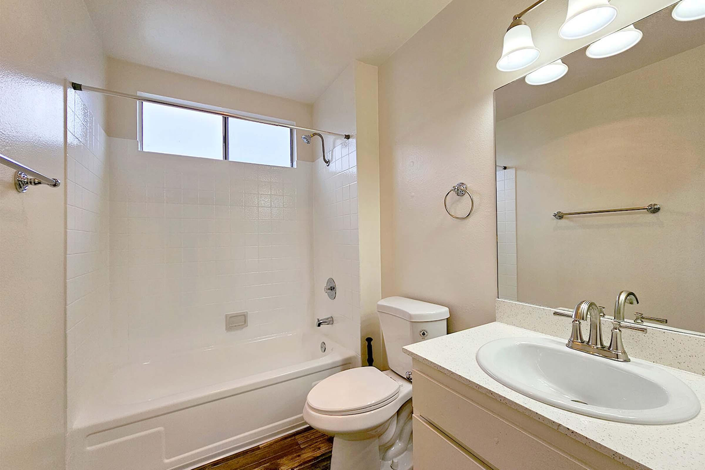 A clean and bright bathroom featuring a white bathtub and shower, a modern sink with a silver faucet, a toilet, and a mirror above the sink. The walls are painted in a light color, and there is a window letting in natural light. The flooring is a warm wood-like material.