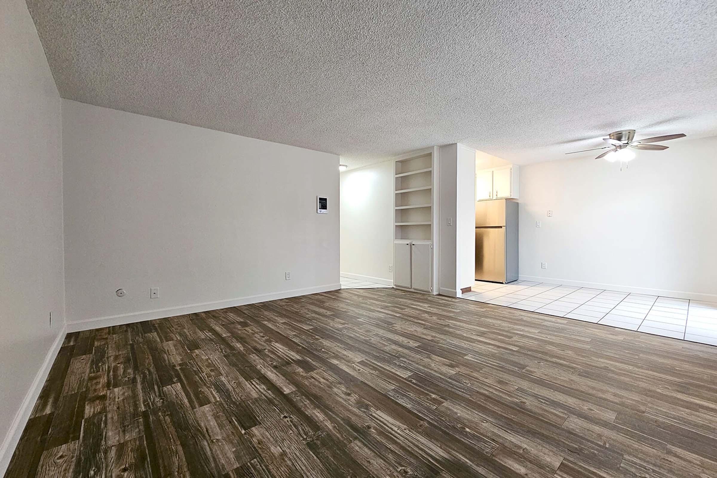 An empty living space featuring wood-style flooring, white walls, and a ceiling fan. On one side, there is a partial wall with built-in shelves. In the background, a tiled area leads to a kitchen, suggesting an open layout. Natural light enters from a nearby window.