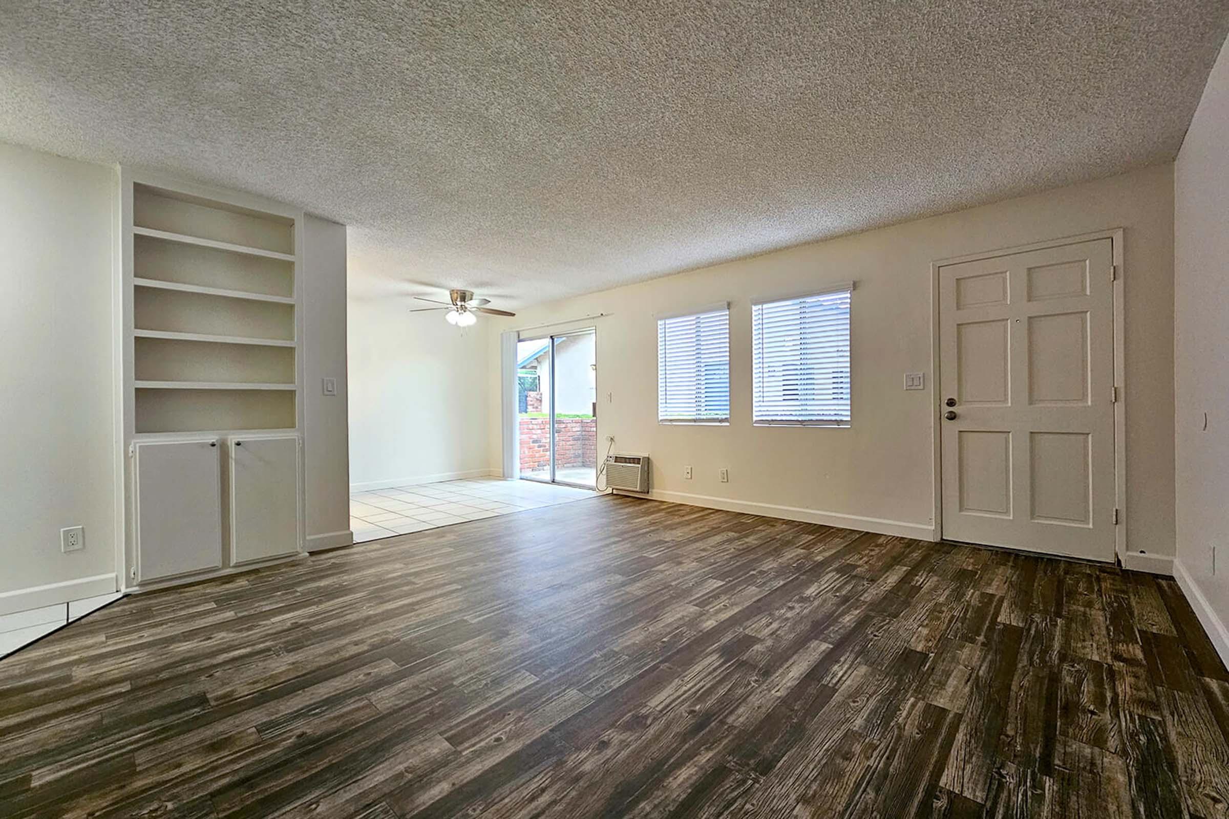 A spacious living area featuring light brown laminate flooring, white walls, and large windows. There is a ceiling fan in the center, built-in shelving on the left, and a door on the right leading to a patio. Natural light fills the room, creating a bright and inviting atmosphere.