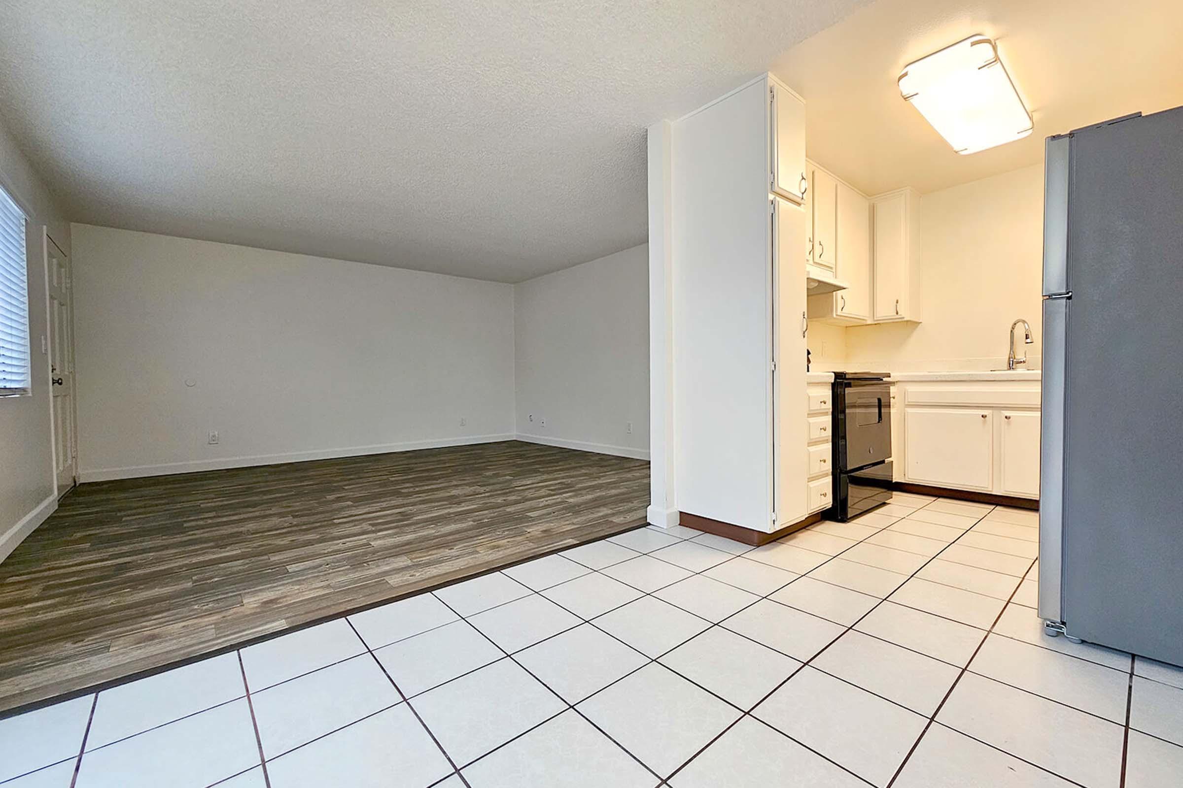 A spacious, open-concept living area featuring light-colored walls and a combination of tile and wood flooring. The kitchen is at the right, with white cabinets, a sink, and a stove. The room is well-lit with a ceiling light, and large windows allow natural light to enter.