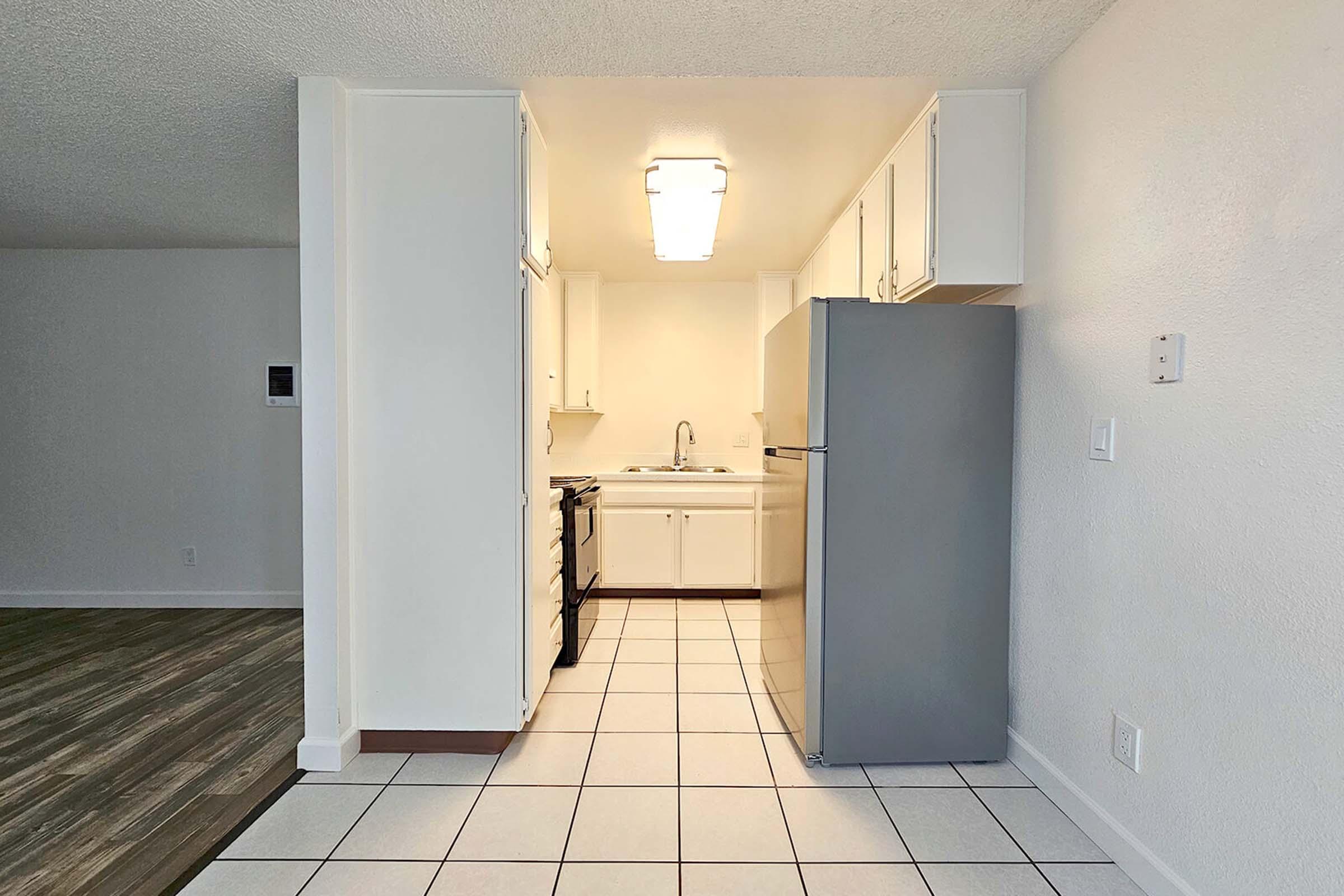 A view of a clean, modern kitchen with white cabinets, a stainless steel refrigerator, and an oven. The kitchen has tiled flooring and is brightly lit by a ceiling light. Adjacent to the kitchen is an open space featuring a wood-look laminate floor. The walls are painted in a light, neutral color.