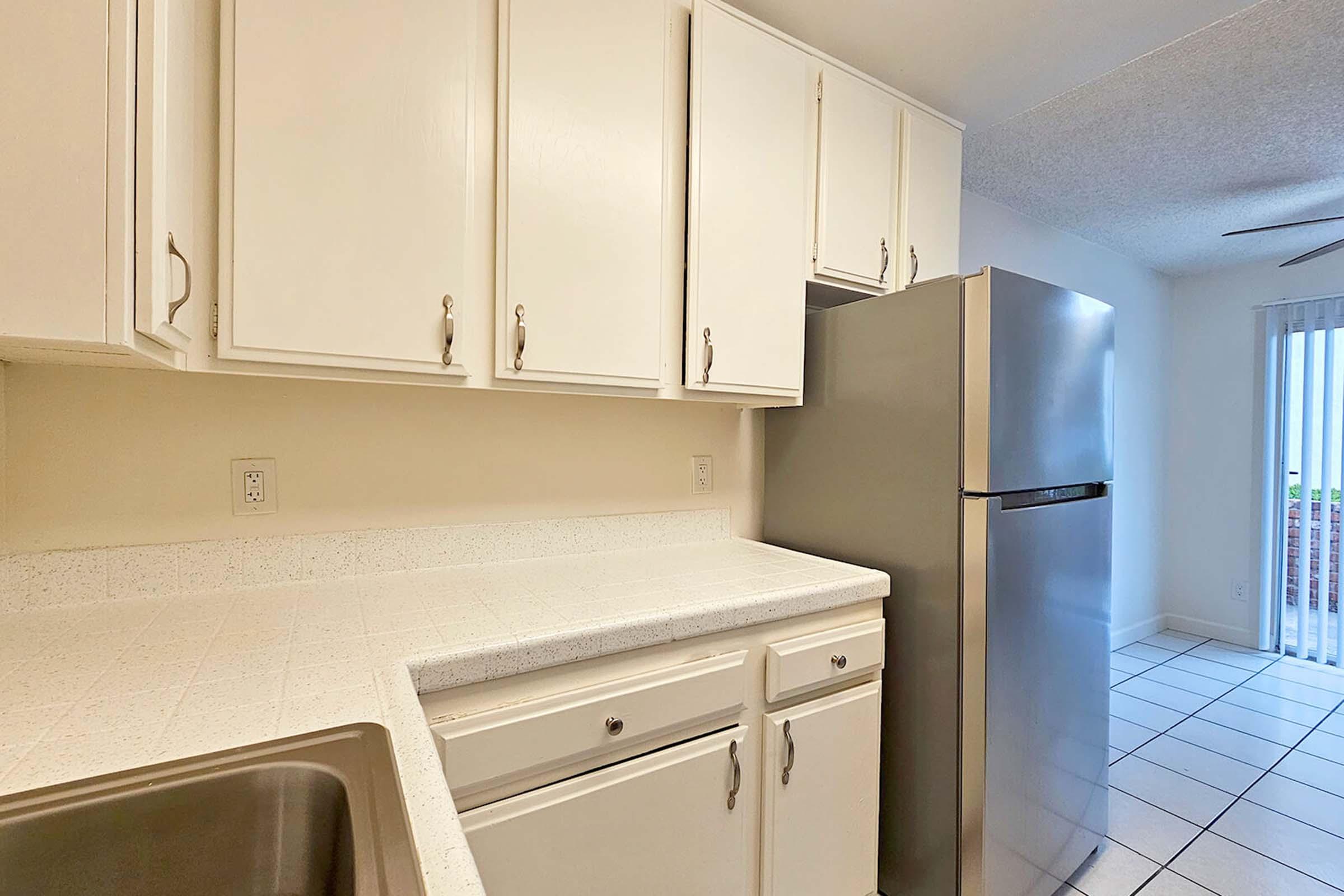 A modern kitchen featuring white cabinets, a stainless steel refrigerator, and a countertop with a sink. The tiled floor and bright lighting create a spacious atmosphere, with a doorway leading to a well-lit area outside.