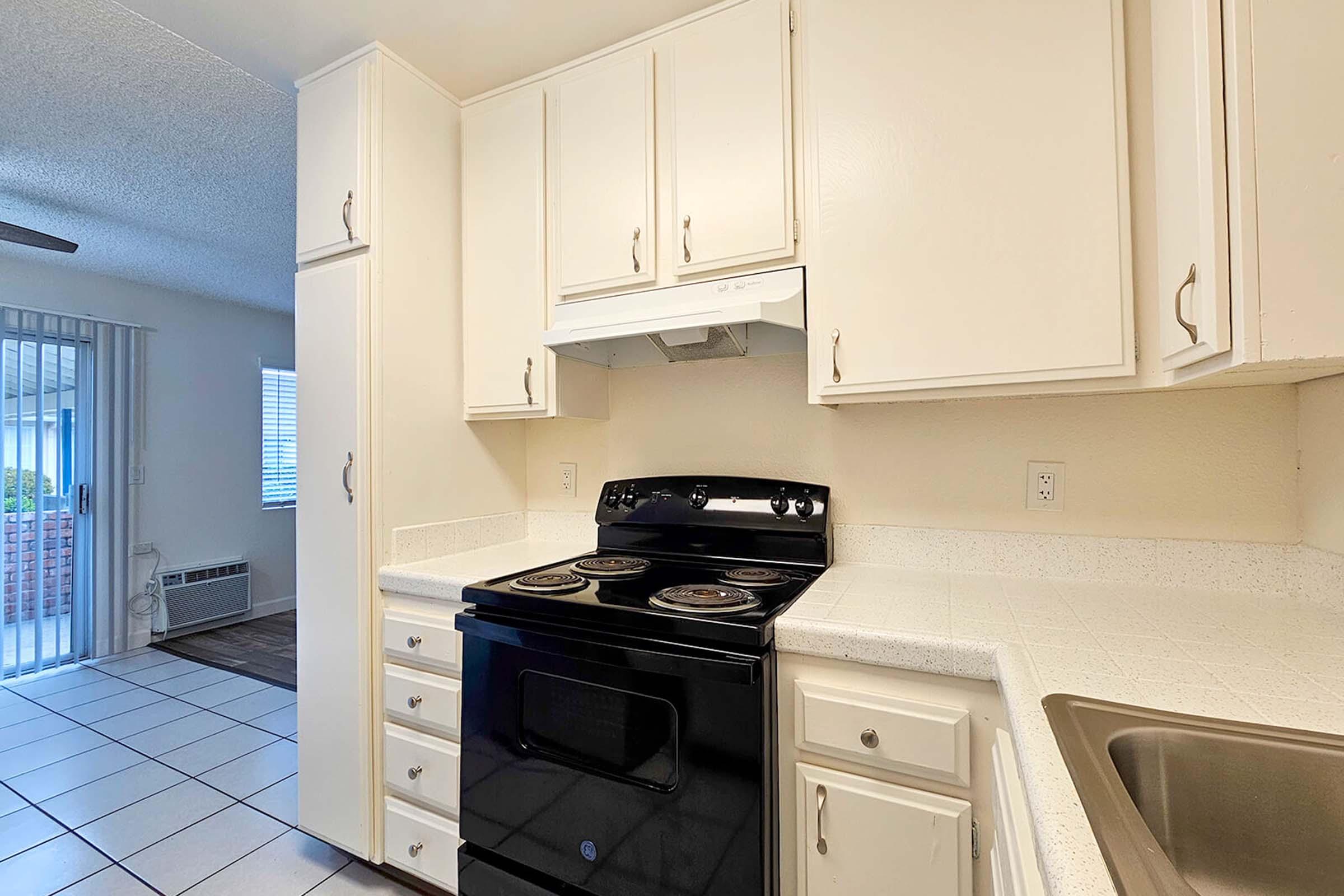 A clean kitchen featuring white cabinets, a black electric stove with four burners, and a kitchen sink. The countertops are light-colored, and there is a window in the background that lets in natural light. The floor is tiled, and there is a small dining area visible through the window.