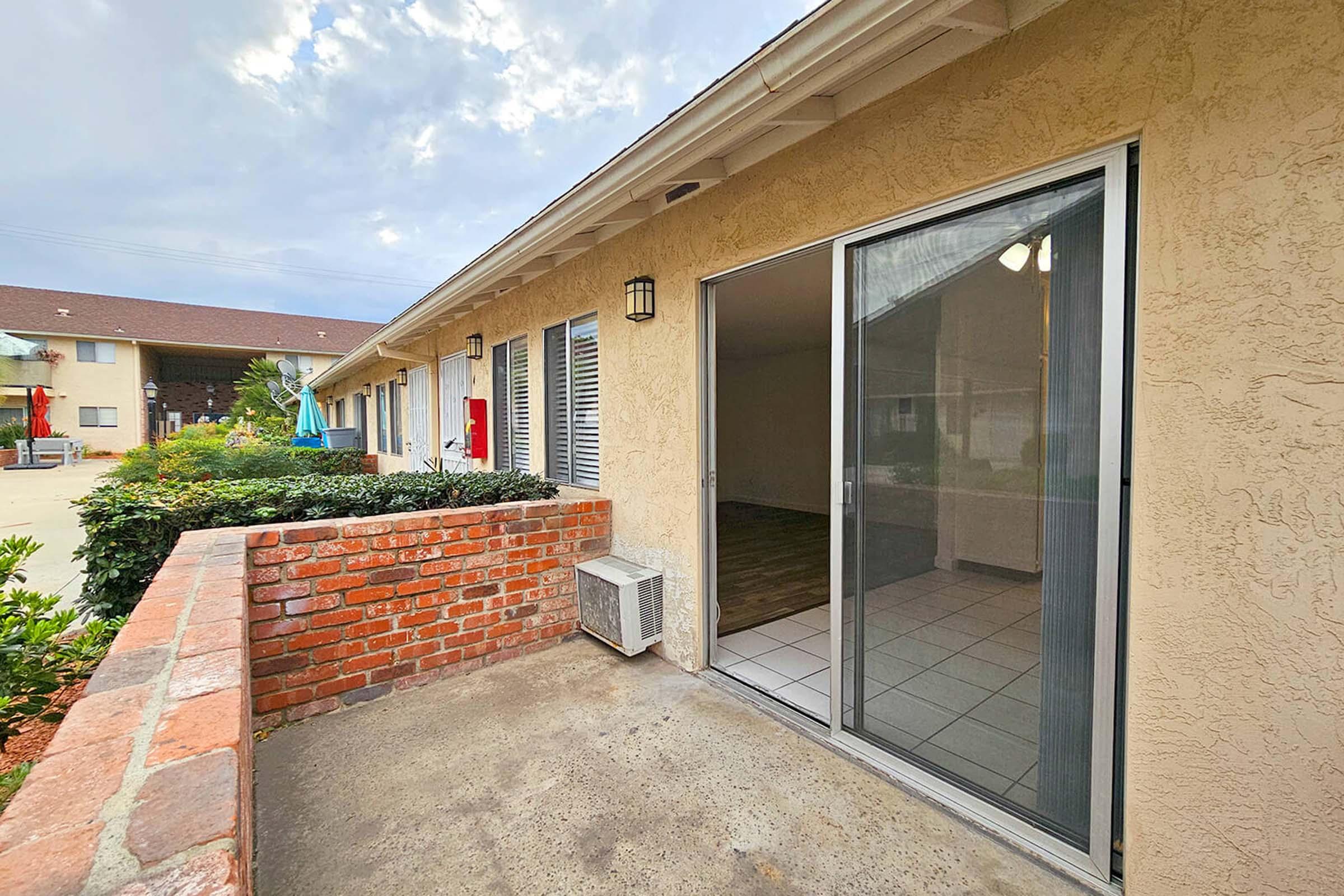 A view of a patio area with a sliding glass door leading into a room. The exterior features a brick wall and an air conditioning unit. In the background, another building is visible, along with some greenery and a cloudy sky.