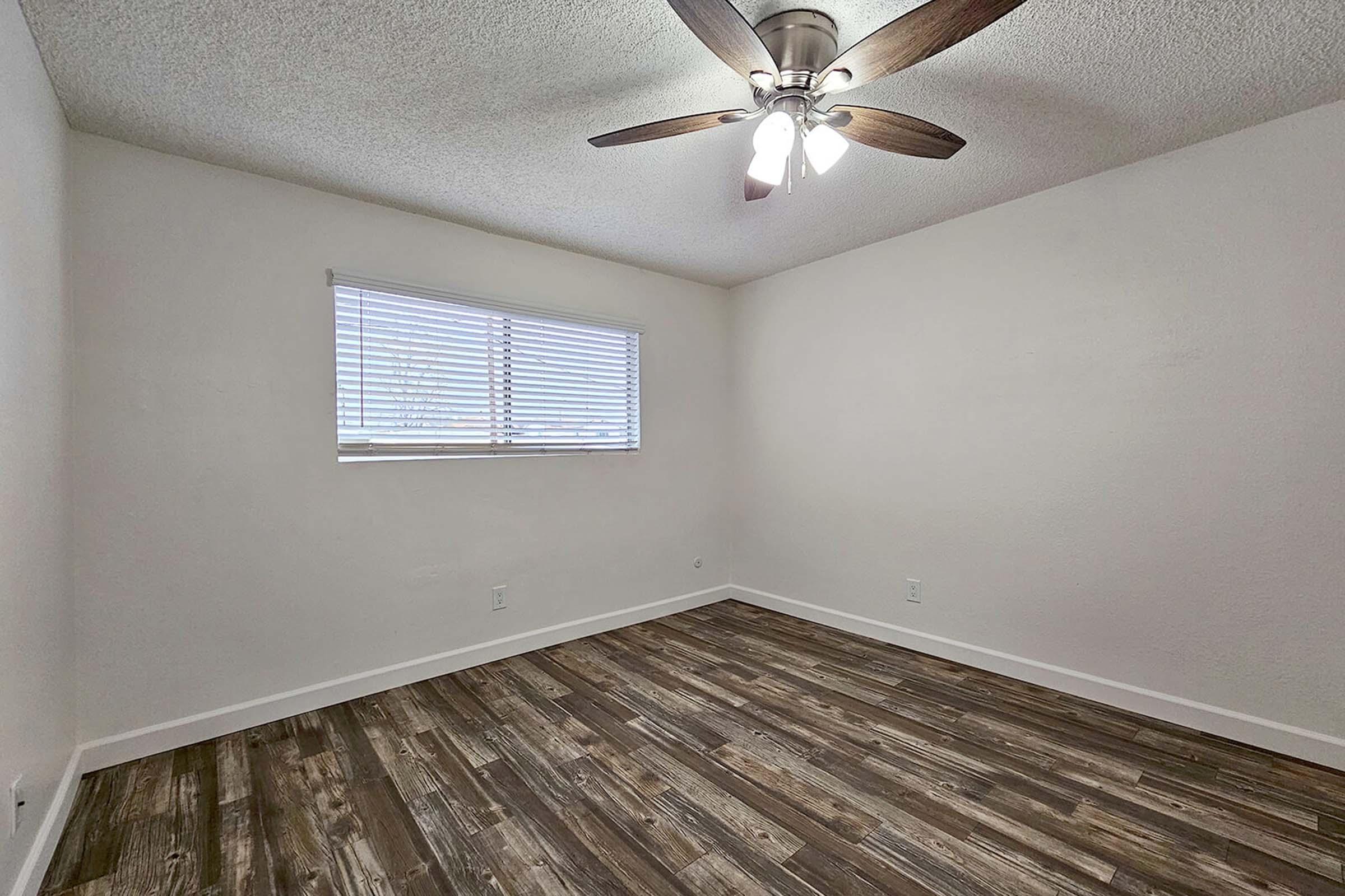 A clean, empty room featuring light-colored walls, a ceiling fan with three lights, and a window with blinds. The floor is covered in wooden laminate planks, creating a warm atmosphere. The space is well-lit and ready for furniture or decoration.