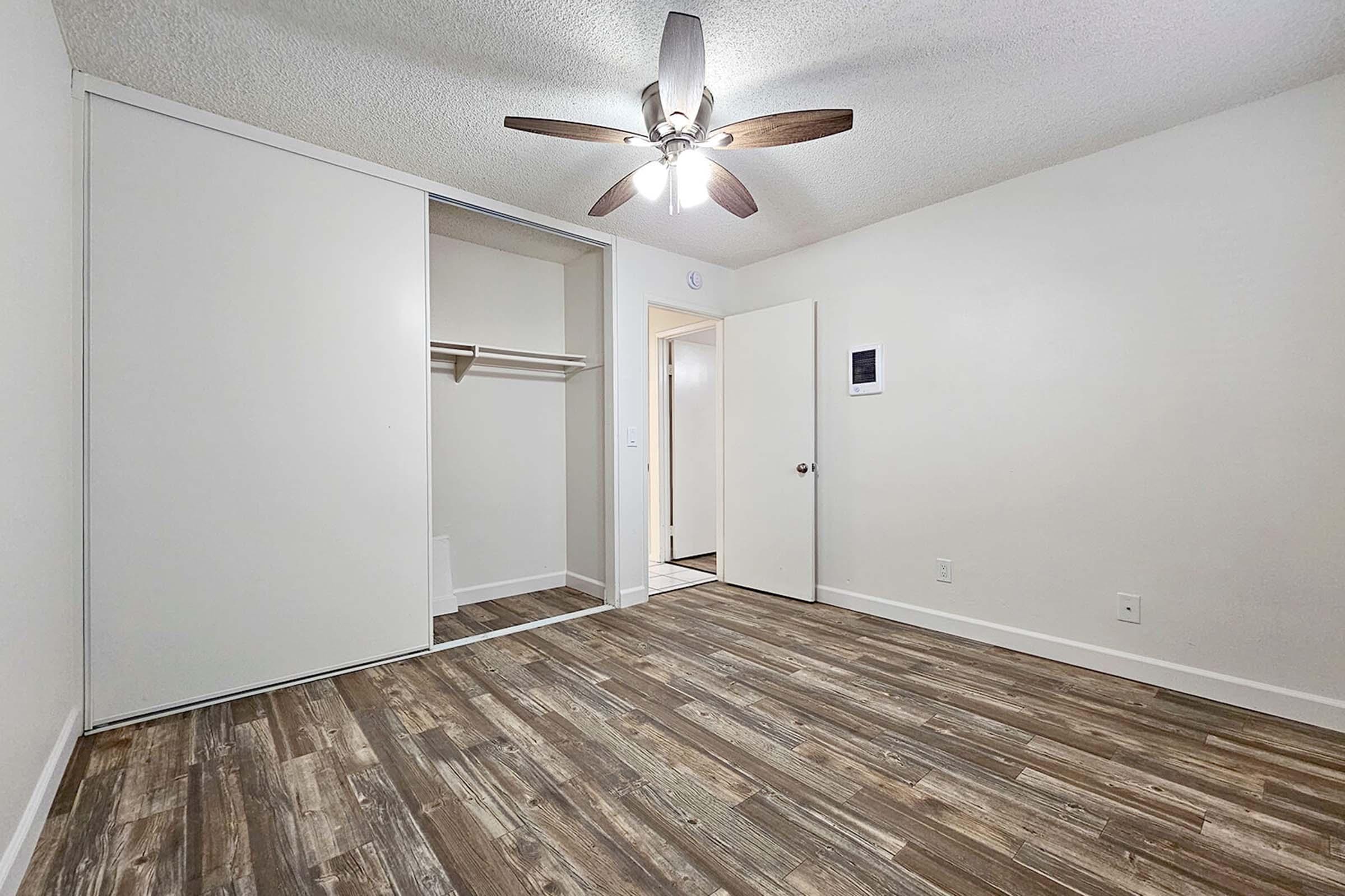 A spacious room featuring a ceiling fan, light-colored walls, and wooden flooring. There is a closet with sliding doors on one side and a doorway leading to another area. The overall look is clean and modern, with natural light enhancing the open feel of the space.