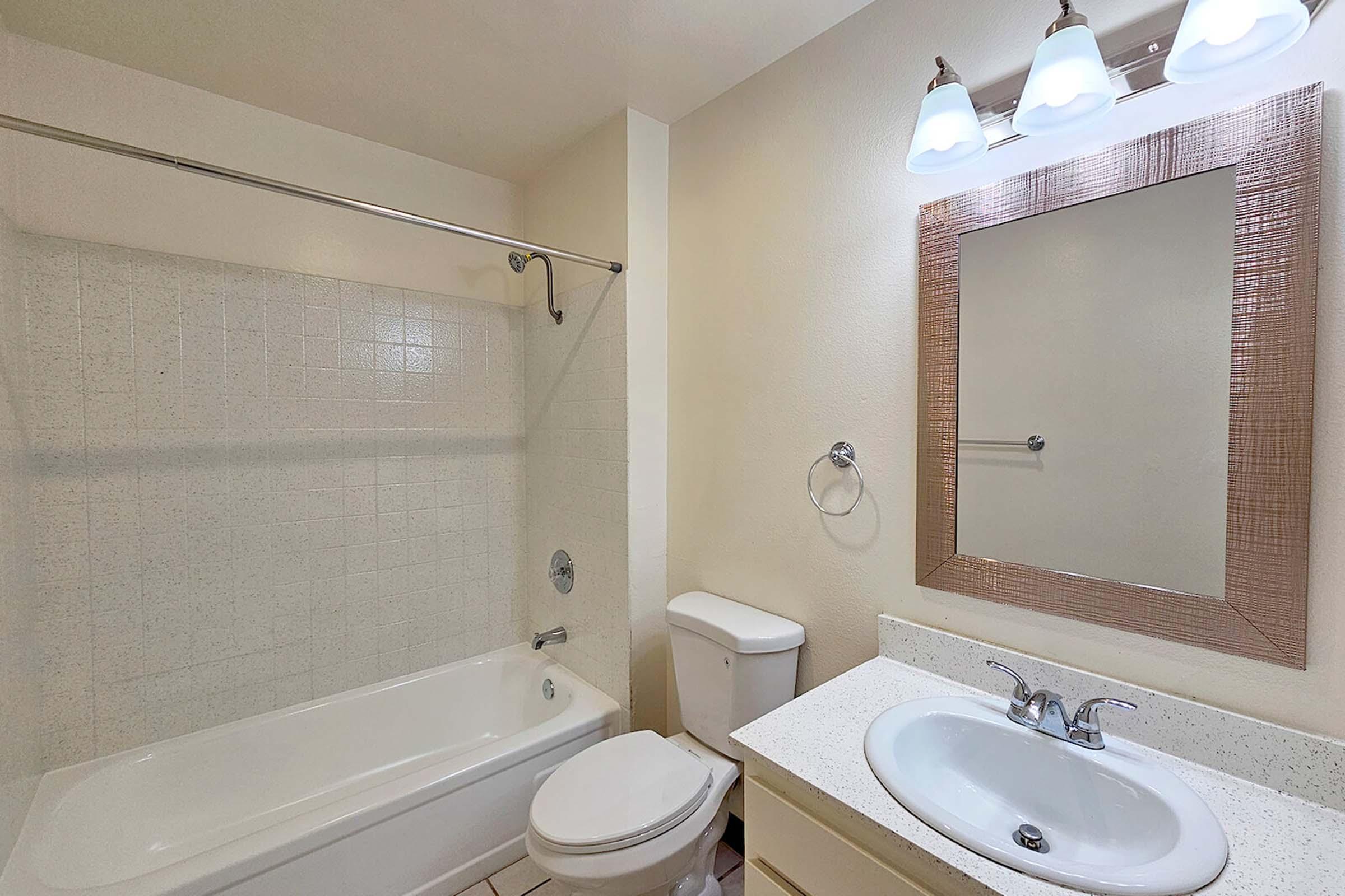A clean and modern bathroom featuring a bathtub with a shower curtain, a white toilet, a sink with a faucet, and a mirror above the sink. Three light fixtures are mounted above the mirror, illuminating the space. The walls are a neutral color, and the flooring is tiled.