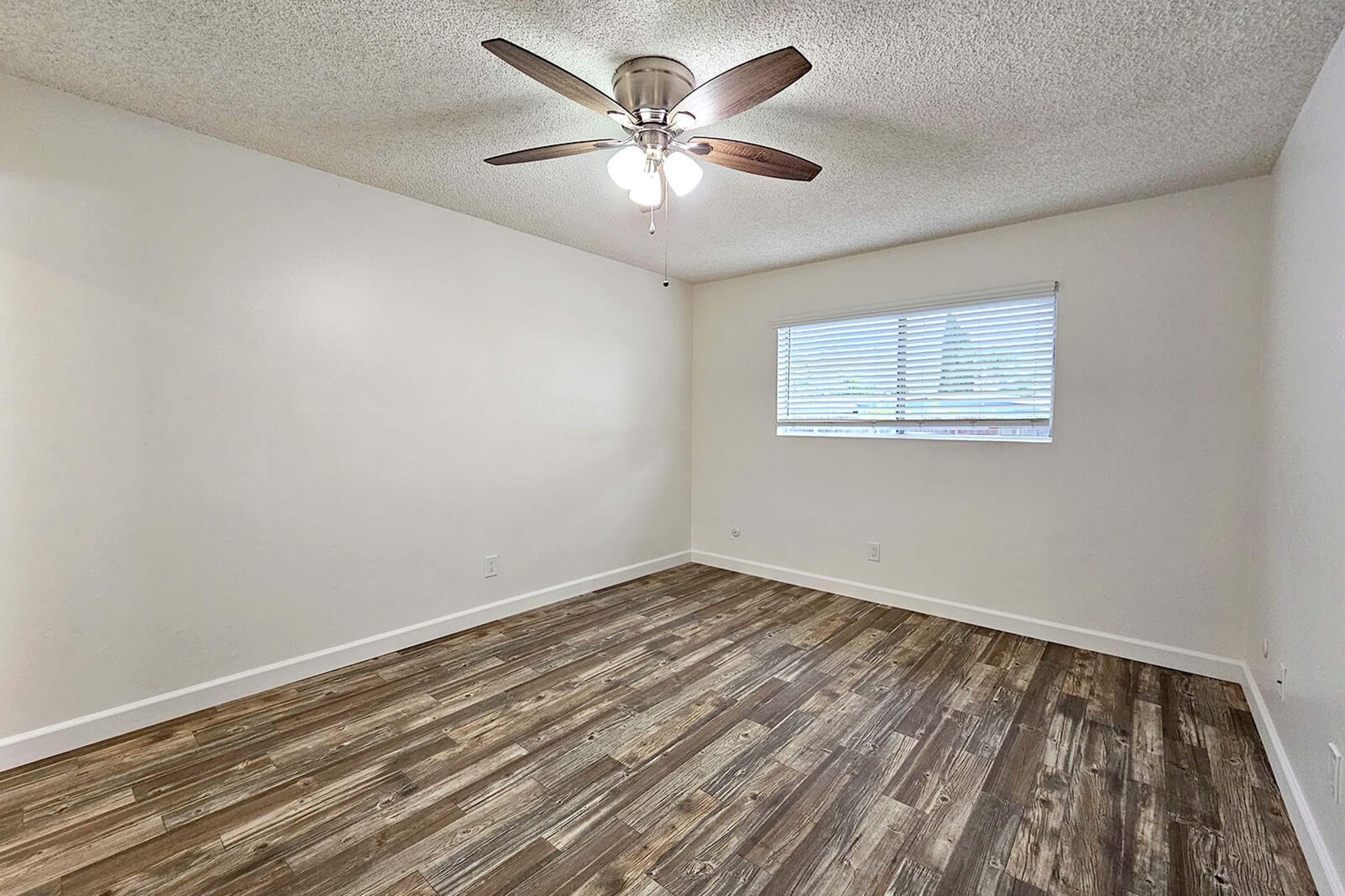 A bright, empty room with a ceiling fan and light fixture. The walls are painted white, and there’s a window with blinds allowing natural light. The floor features wood-like laminate flooring, enhancing the spacious feel of the room. Suitable for various purposes such as a bedroom or office.