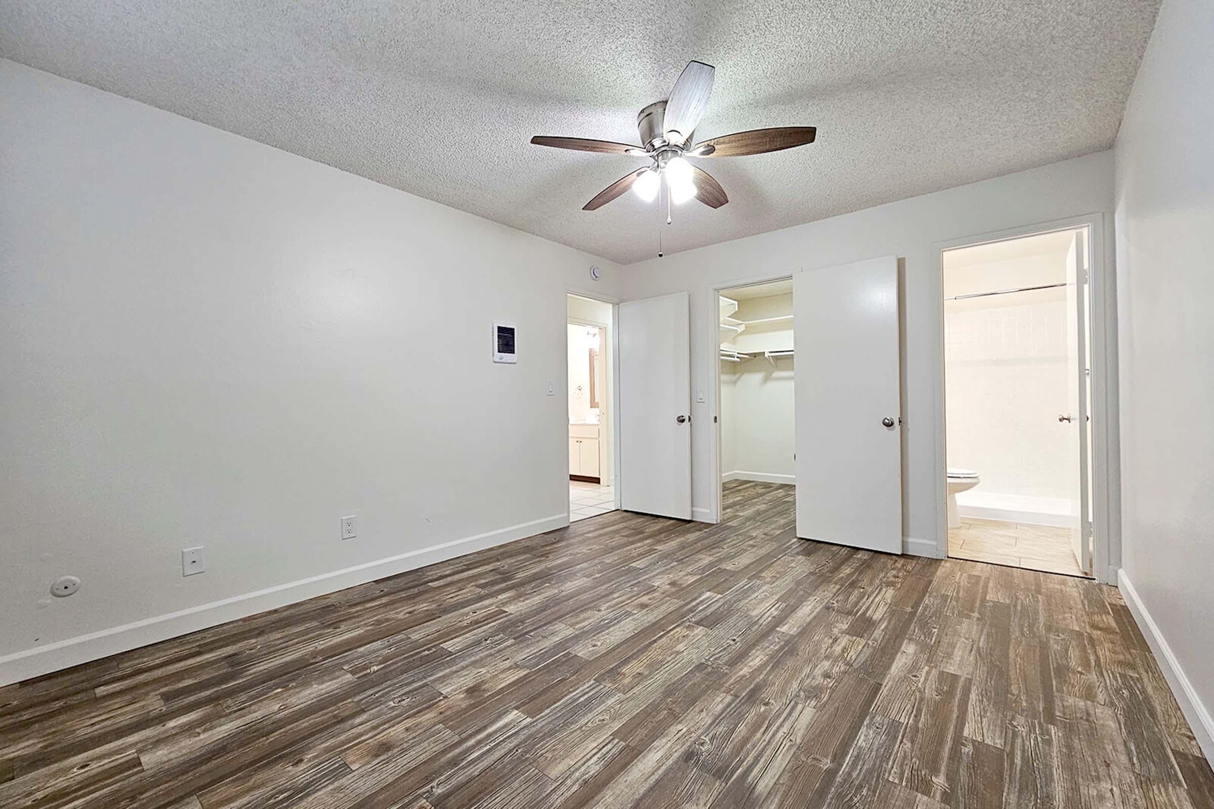 A spacious room featuring light-colored walls and a ceiling fan, with a wooden floor. Two closed doors lead to adjoining spaces, one likely containing a closet. Natural light is present, enhancing the neutral decor. The overall atmosphere is bright and inviting, suitable for various uses.