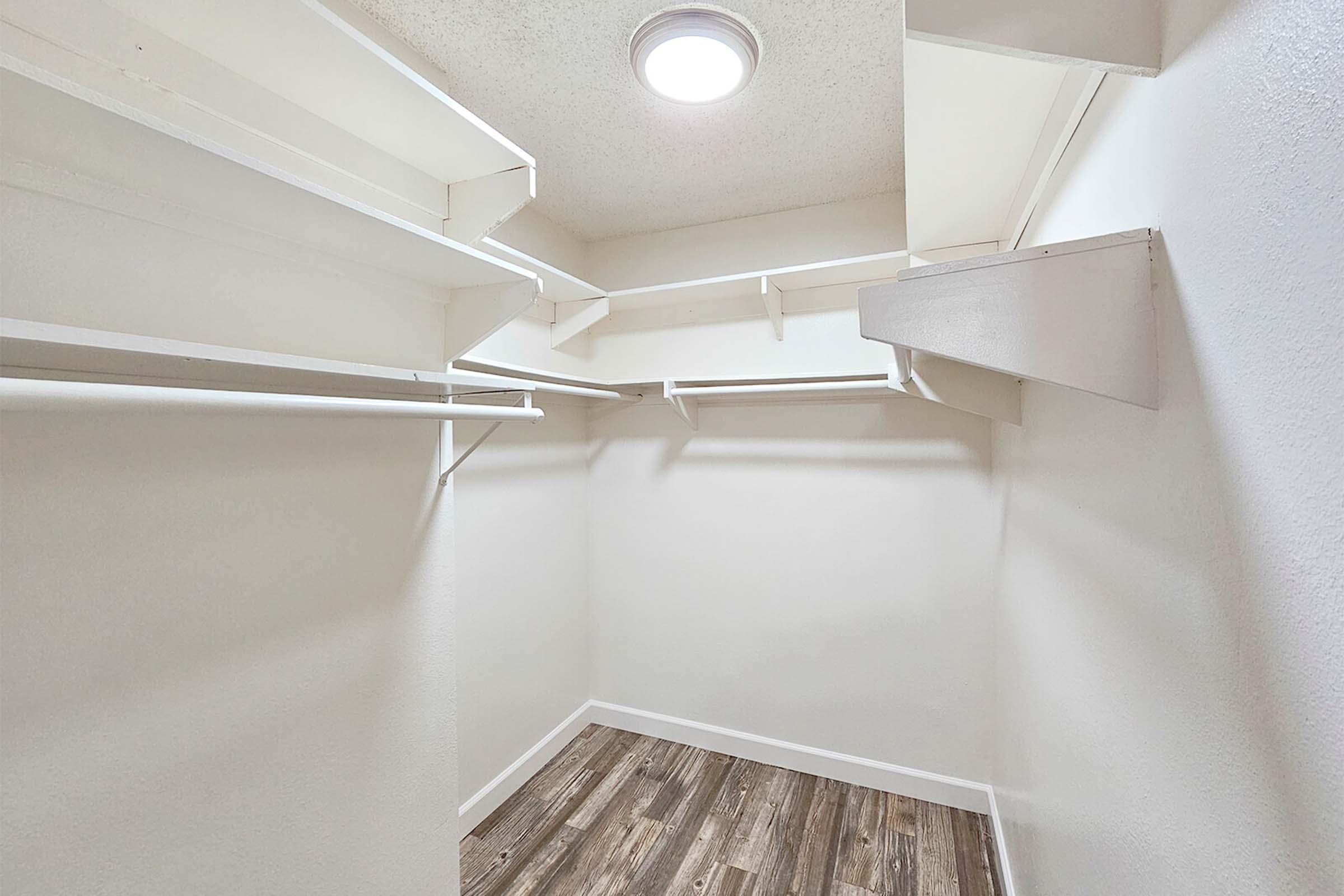 Empty closet with white walls and several shelving units installed. The floor is covered with laminate planks, and a ceiling light fixture illuminates the space. The closet appears spacious and organized, ideal for storage.