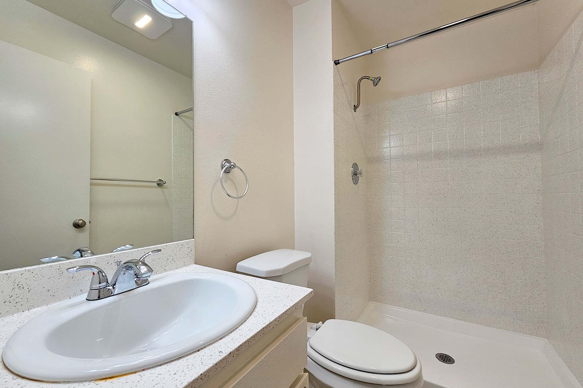 A bathroom featuring a white sink with two faucets, a toilet, and a shower area with a glass sliding door. The walls are painted a light color, and a mirror is mounted above the sink. There is a towel ring on the wall, and the overall design is simple and modern.