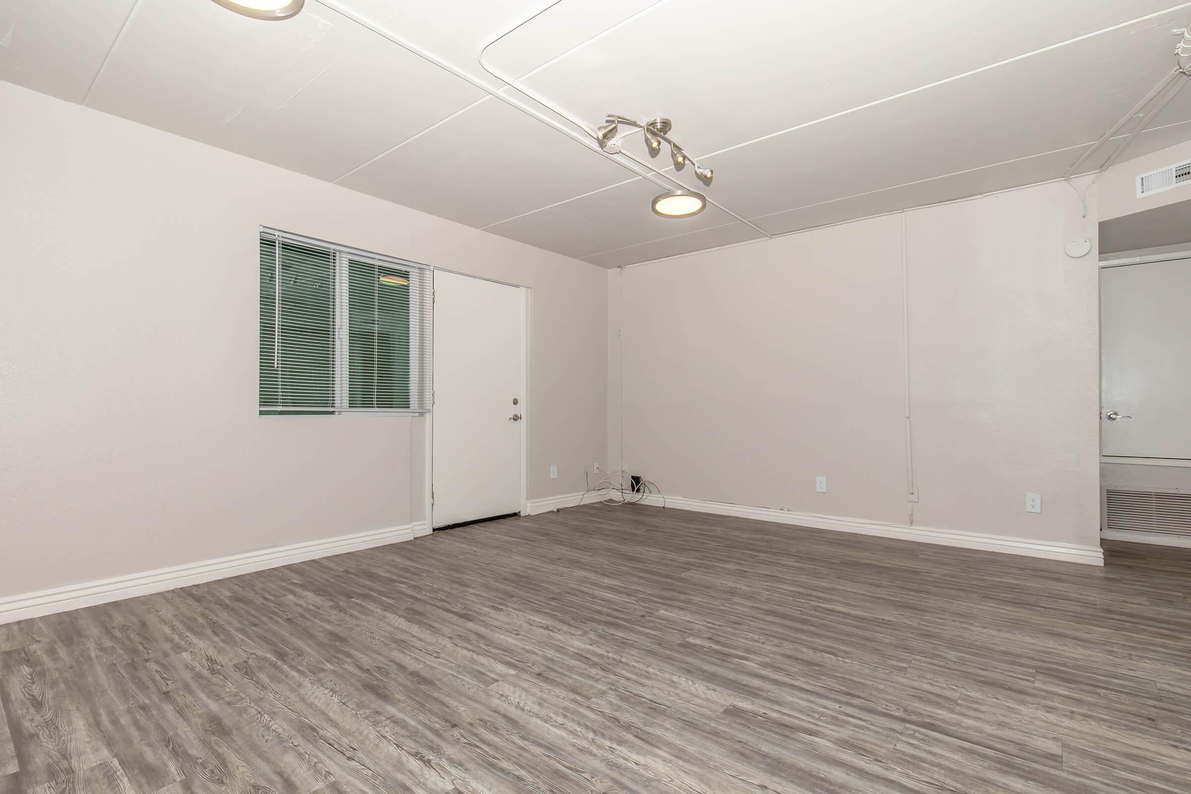Empty interior of a room featuring light-colored walls, a window, and a door. The floor has a wood-like finish, and there are no furniture or decorations in the space. Ceiling lights are present and the overall atmosphere is bright and clean.