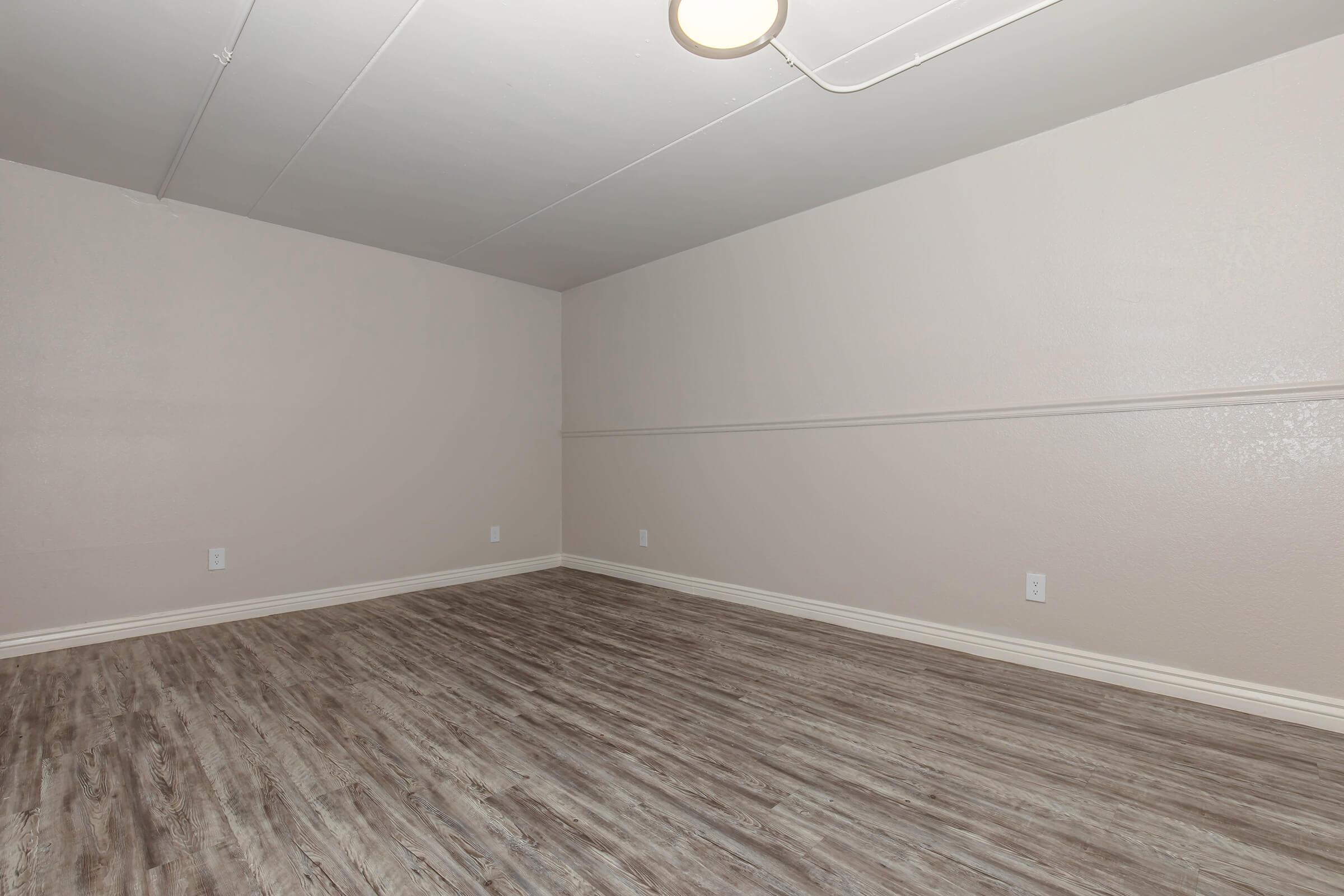 A vacant room with light beige walls and a wooden floor. The ceiling has a circular light fixture, and there are no furniture or decorations present. The space appears clean and well-maintained, with baseboards along the lower wall.