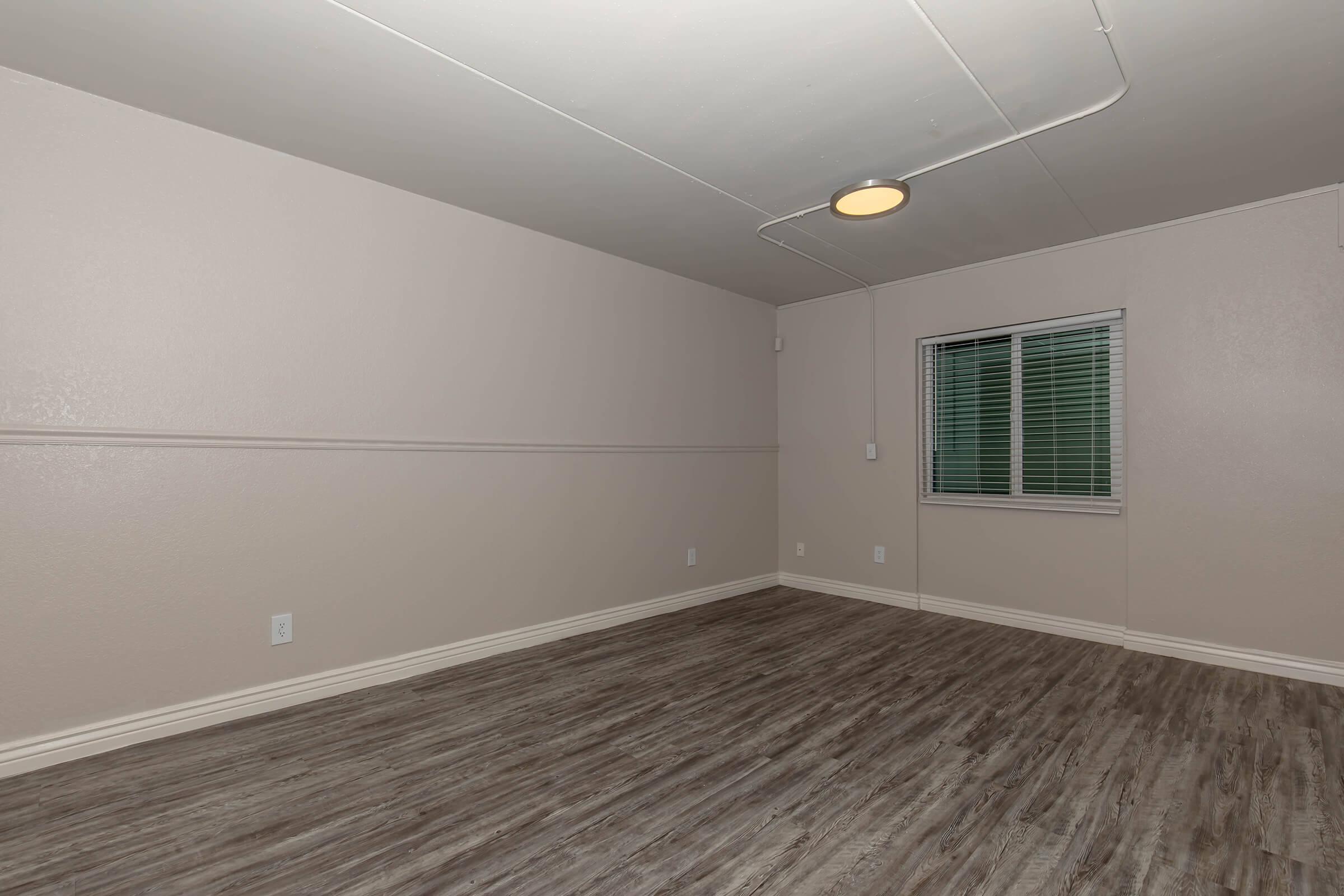 Empty room with light-colored, neutral walls and a wooden floor. A small window with blinds lets in natural light. The ceiling features a simple light fixture. The space appears clean and unfurnished, creating a minimalistic atmosphere.