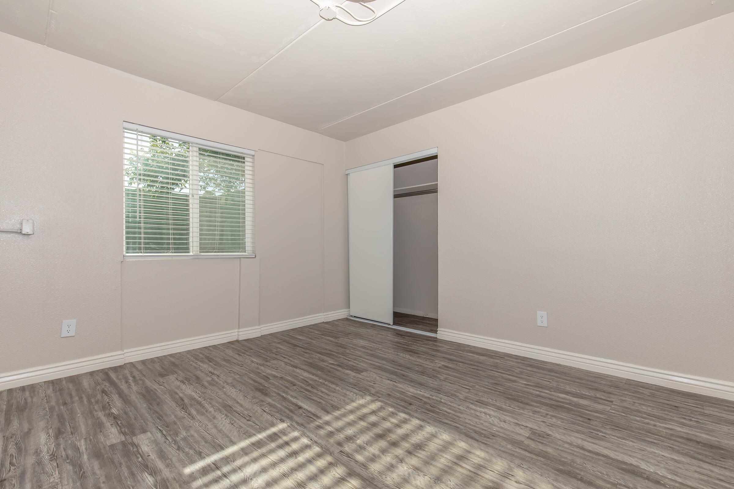 An empty room with light-colored walls and a wooden floor. There is a window with blinds allowing natural light to enter, and a closet with sliding doors. The room has no furniture, creating a spacious and open feel.