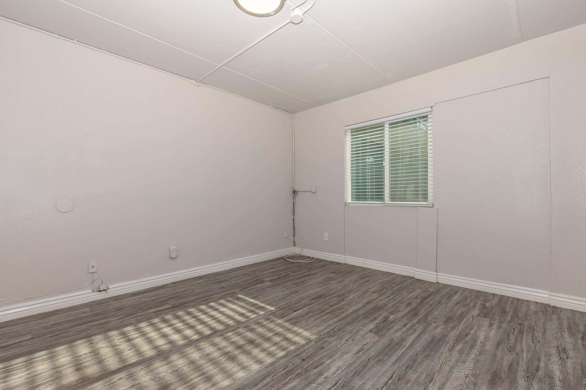 Empty room with light-colored walls and natural wood laminate flooring. A window with blinds is situated on one wall, allowing natural light to enter. There's minimal furniture and no decor, creating a spacious and clean atmosphere.