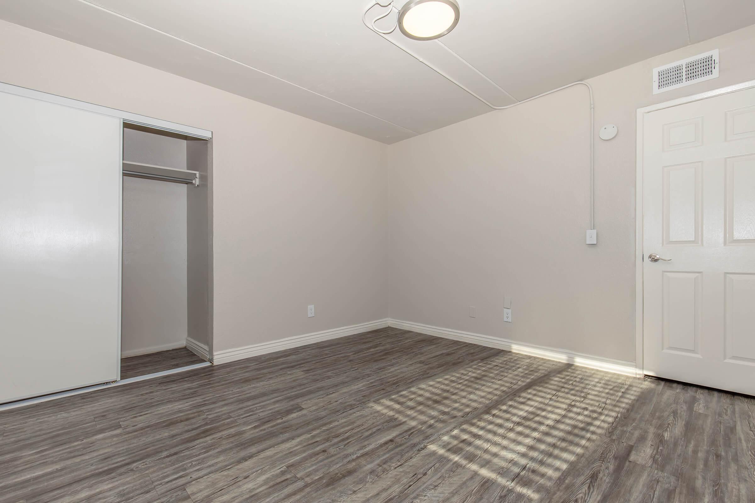 Empty room with light-colored walls and a wooden floor. A white closet is visible on the left, and there is a door on the right. The room has a ceiling light and is spacious with no furniture present, creating a minimalist appearance. Shadows from the light source can be seen on the floor.