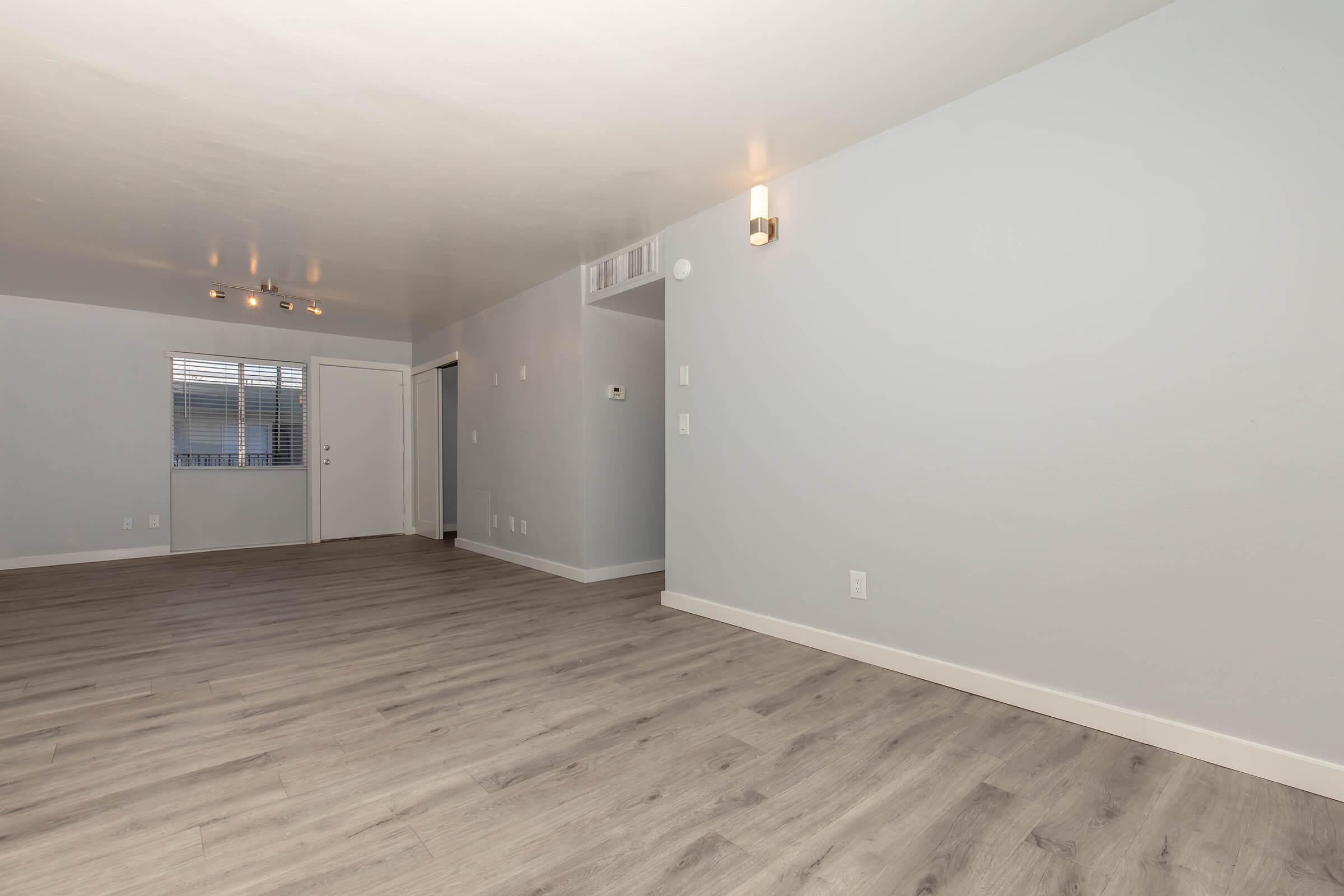 Empty room with light-colored walls and a smooth wooden floor. It features a window on one side, a door leading outside, and a wall-mounted light fixture. The space is well-lit, creating a clean and modern atmosphere.
