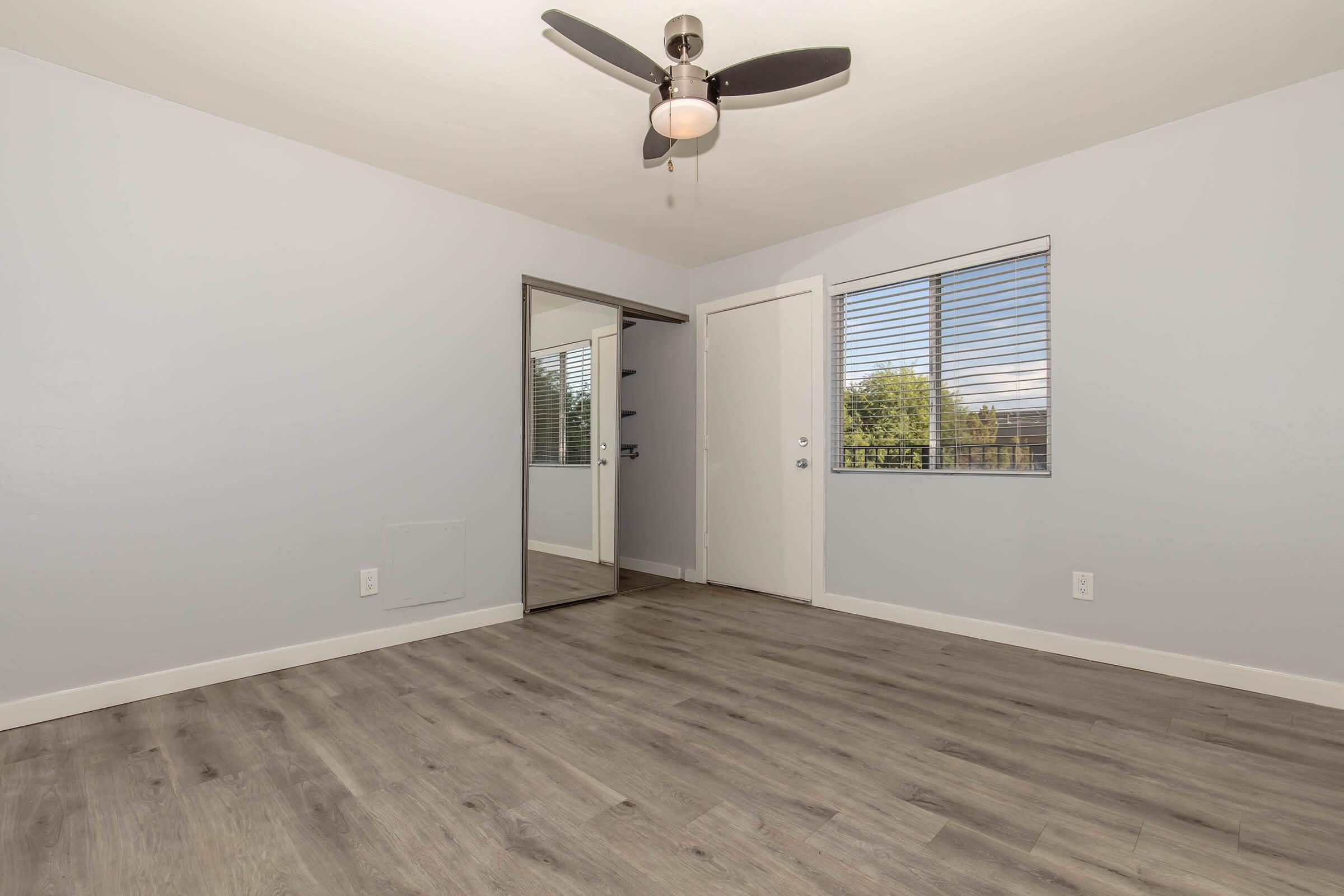 A spacious room with light gray walls and wooden flooring. It features a ceiling fan, a large mirror on the wall, and a window with blinds allowing natural light to enter. A door leads to another area, creating a bright and airy atmosphere.