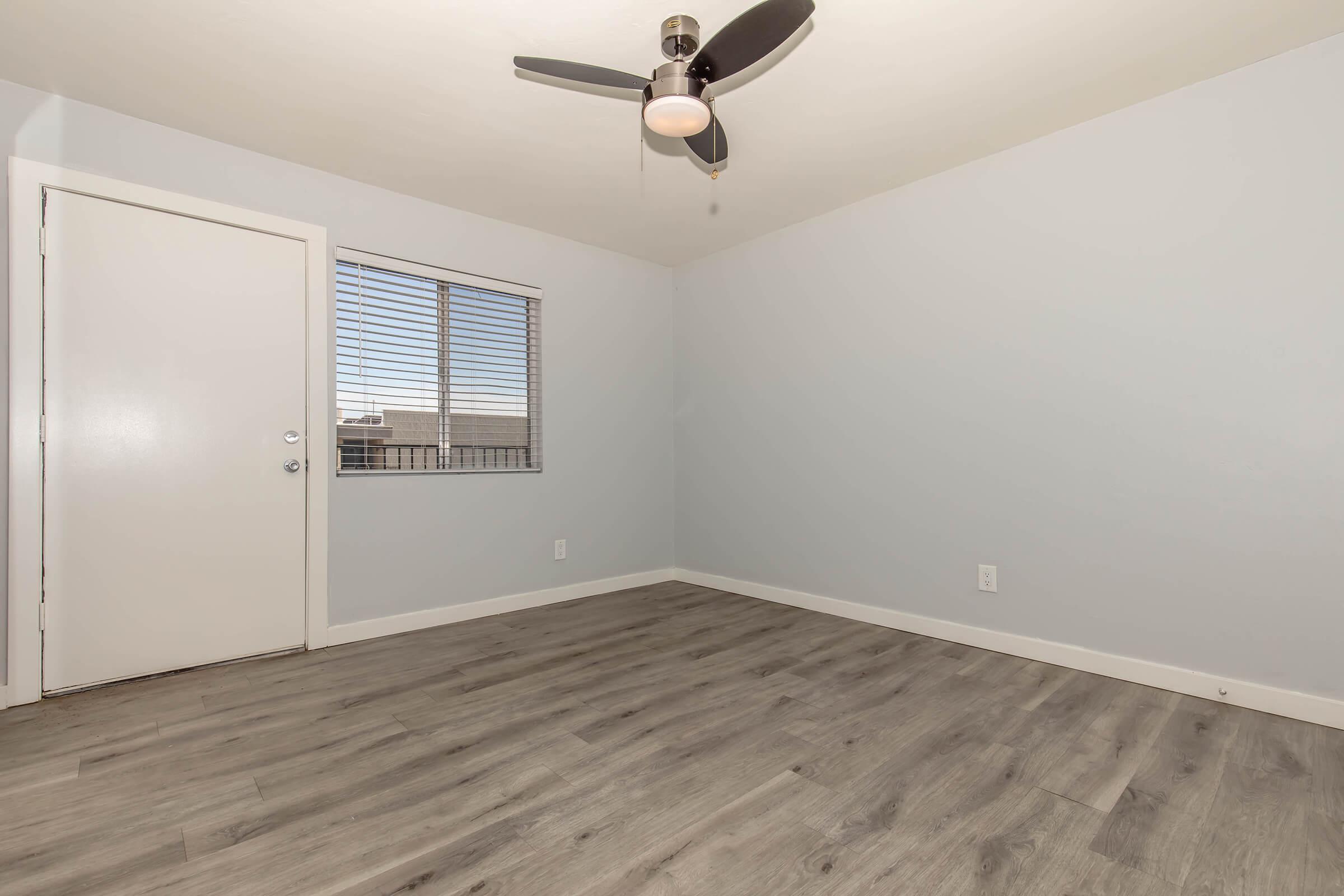 A minimalistic room featuring light gray walls and a ceiling fan. There is a window with blinds on one wall allowing natural light, and a simple, closed door on the adjacent wall. The floor is a light wood-like laminate, giving the space a modern and clean appearance.