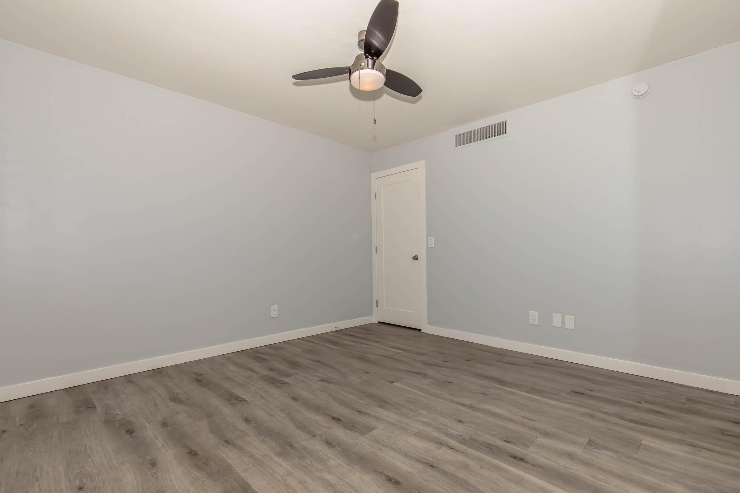 Empty room featuring light gray walls, new laminate flooring, and a ceiling fan. A single door is visible on the right. The space is well-lit with natural light, creating a clean and modern atmosphere. Ideal for personal customization or as a minimalistic living area.