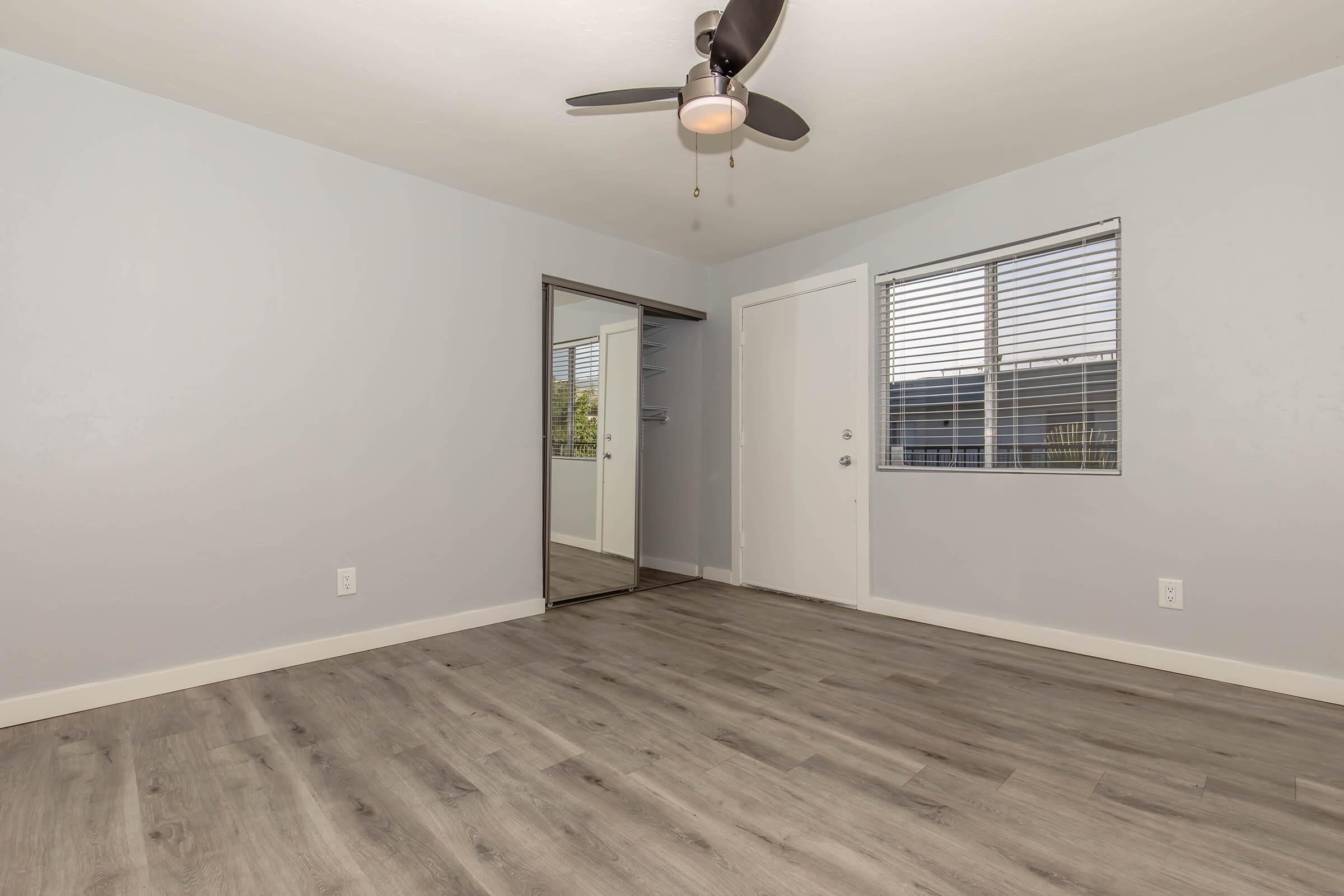 A spacious room with light gray walls and wooden flooring. It features a ceiling fan, a large mirror closet, and a window with blinds. The room is empty, giving a bright and airy feel, ideal for various uses. A door on the right leads to another area.
