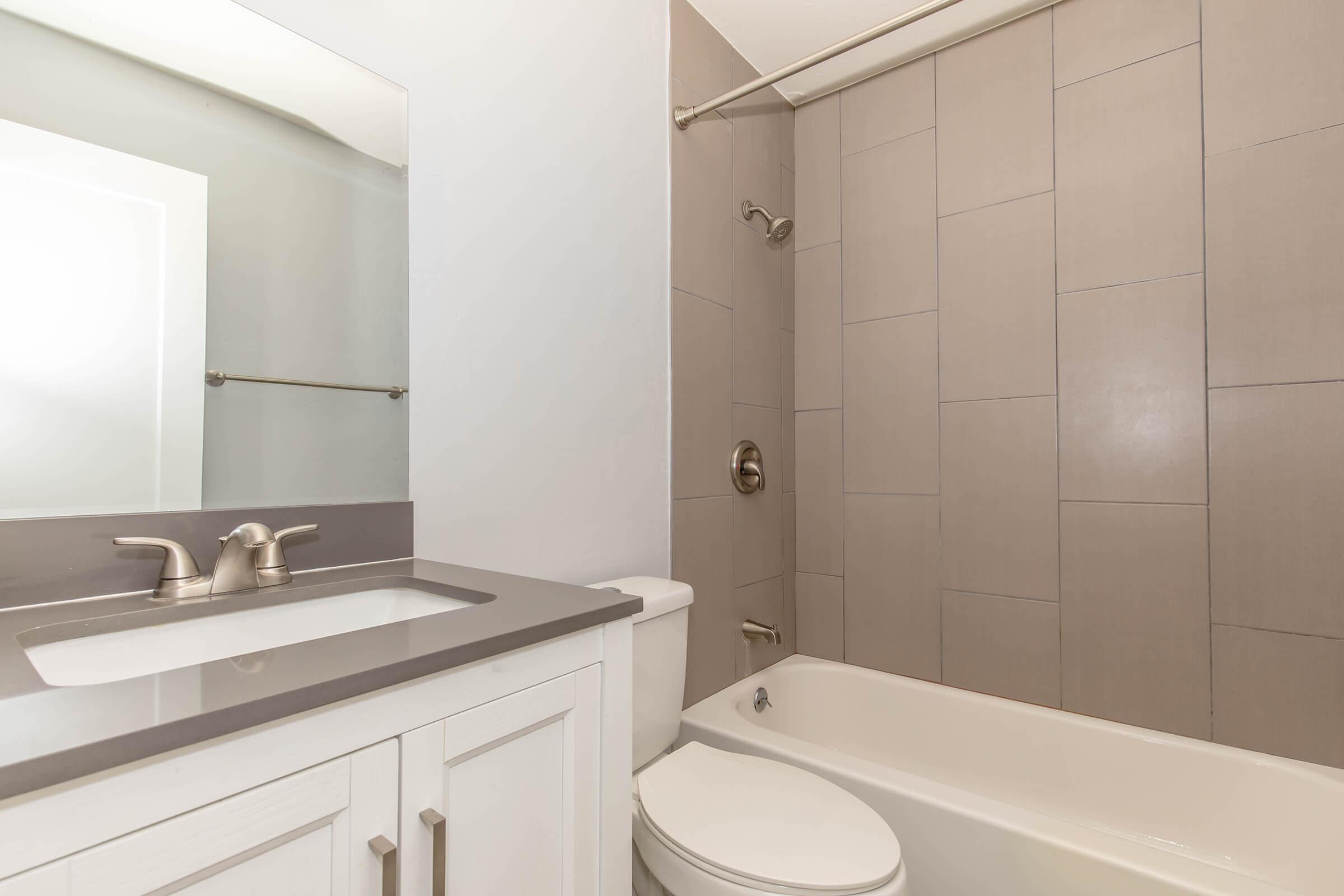 A modern bathroom featuring a white vanity with a sink, a bathtub with a shower, gray tiled walls, and a mirror. The fixtures are silver, and the overall design is clean and contemporary.