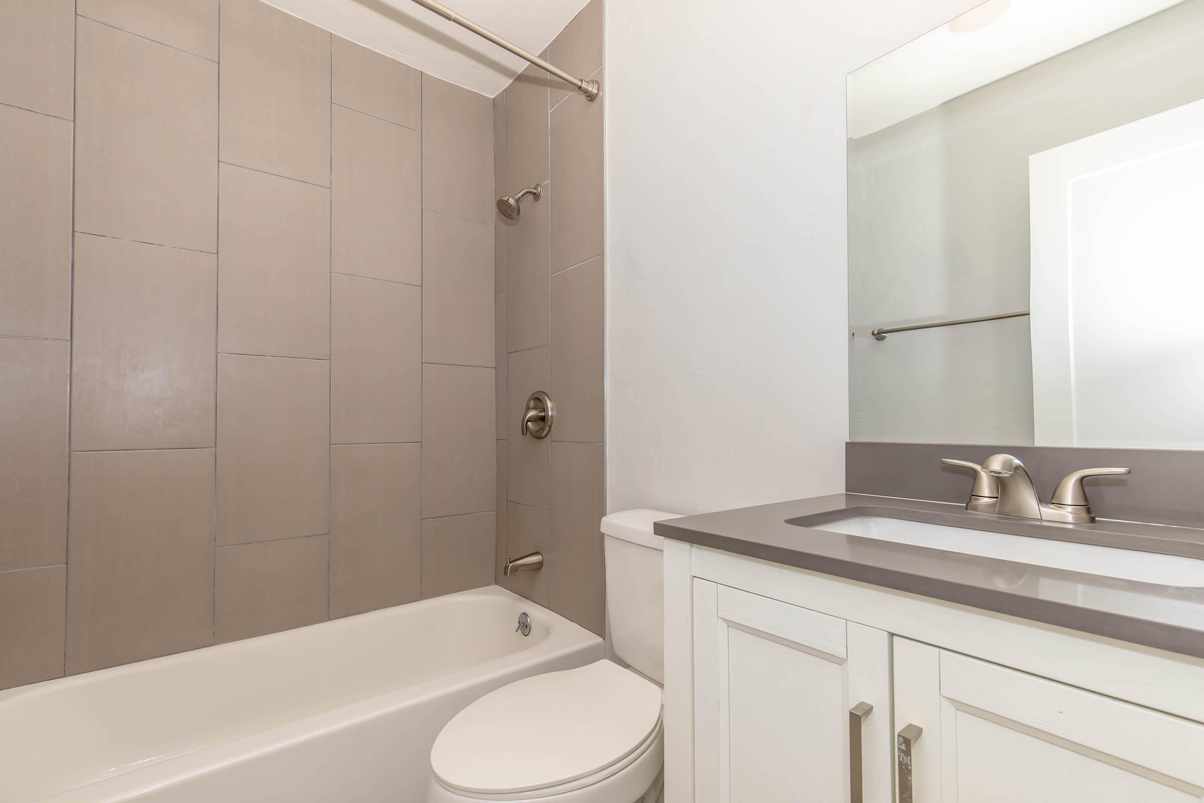 A clean, modern bathroom featuring a shower-tub combination with tiled walls, a white vanity with a gray countertop, and a large mirror. The space is well-lit, showcasing neutral colors and contemporary fixtures.