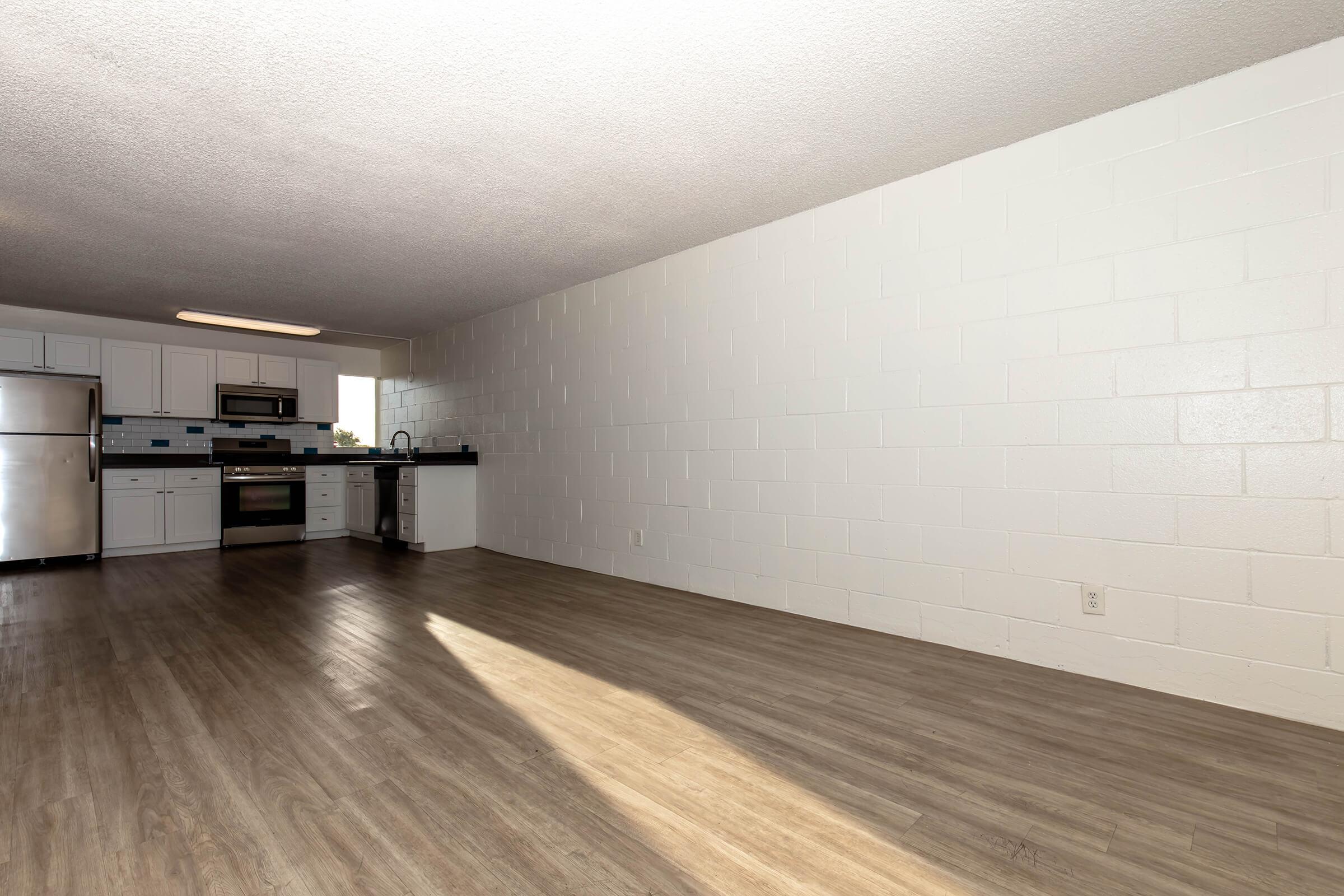 A spacious, empty room featuring a modern kitchen with stainless steel appliances. The kitchen has white walls, a black countertop, and a window. The floor is a light wood laminate, and there is ample natural light illuminating the space.