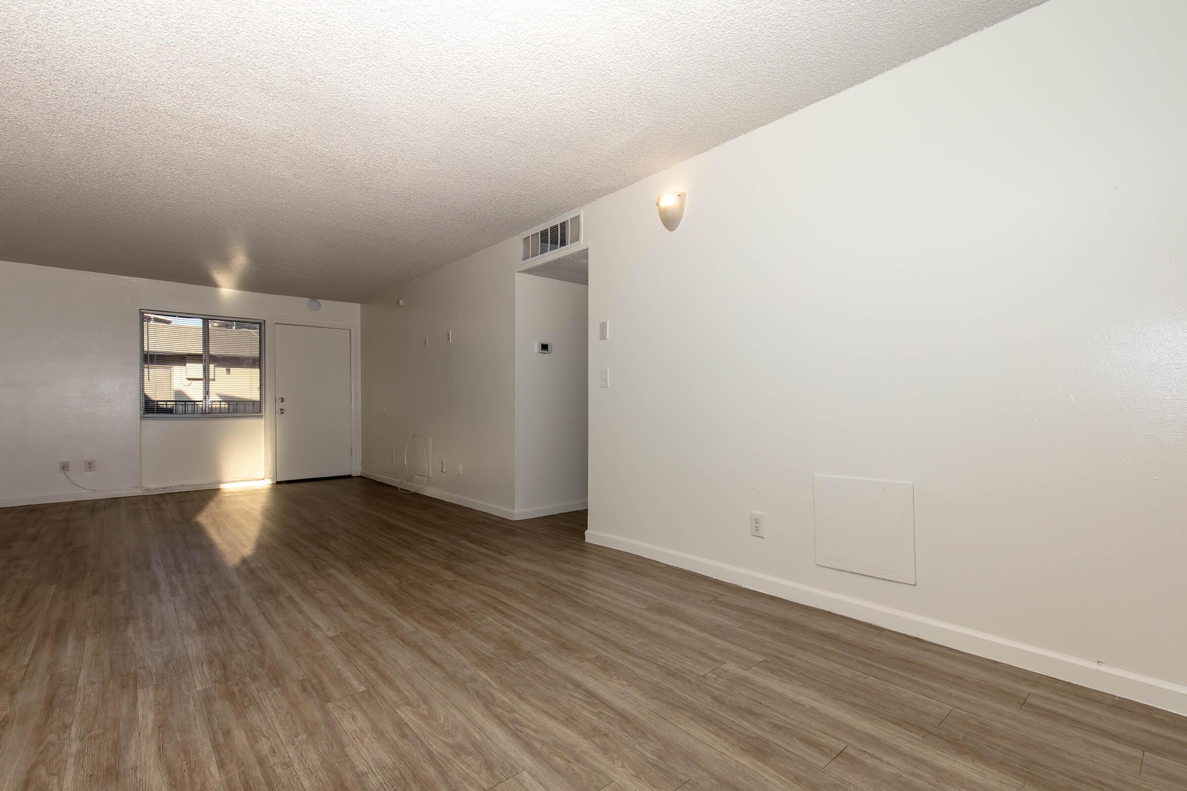 Spacious empty room with light wood-like flooring, white walls, and a small window. Features a door on one wall, providing natural light. The ceiling is textured, and there is a light fixture mounted on the wall. The overall ambiance is bright and inviting, suitable for various interior layouts and designs.